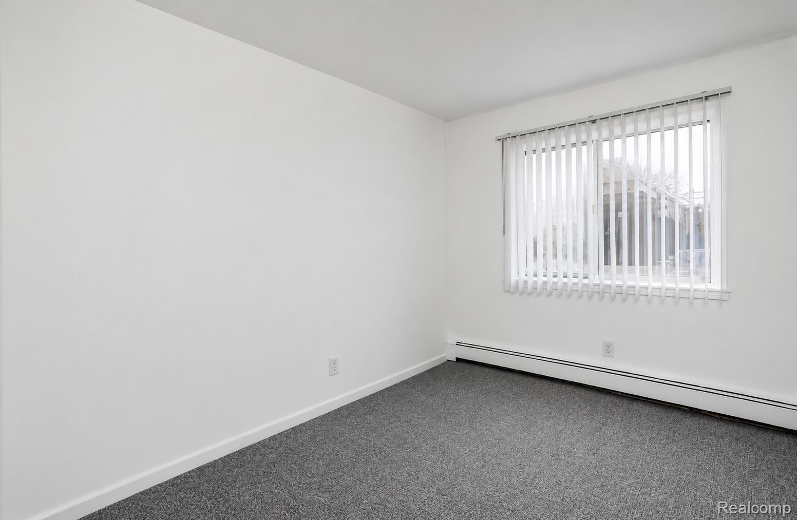 White-painted walls with a window featuring vertical blinds, gray carpet flooring, and baseboard heating
