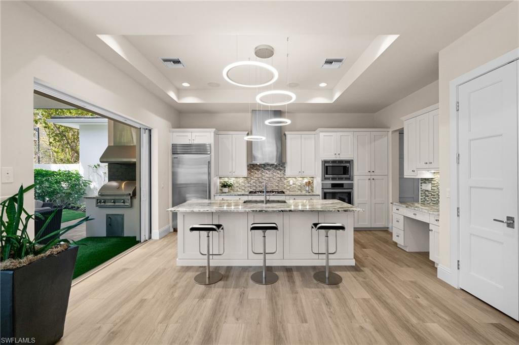 9172 Mercato Lane Naples, FL 34108 - Photo 10 of 29 Kitchen with a raised ceiling, light stone counters, tasteful backsplash, white cabinets, and an island with sink