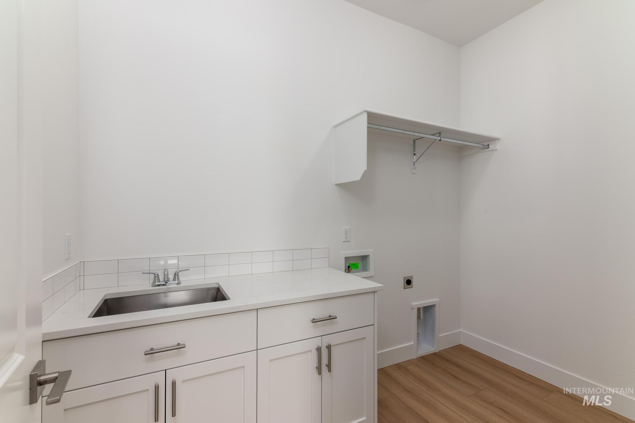 1195 West Switchgrass Drive Meridian, ID 83642 - Photo 11 of 22 Laundry room featuring a stainless steel sink, white cabinetry with brushed nickel hardware, light-toned countertop, white subway tile backsplash, and wood-finish flooring