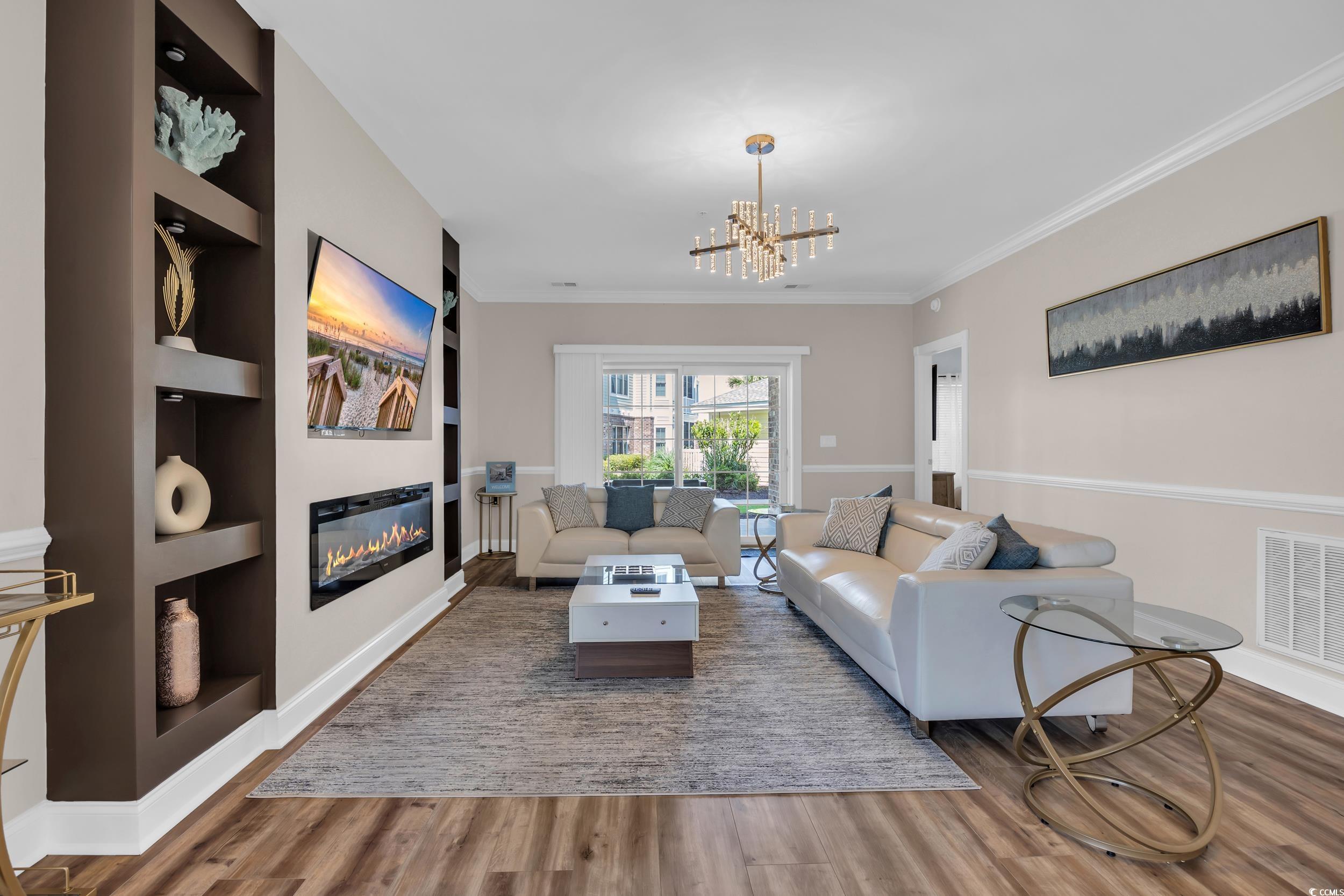 4821 Luster Leaf Circle, Unit 101 Myrtle Beach, SC 29577 - Photo 23 of 30 Living room with built in shelves, a glass covered fireplace, wood finished floors, crown molding, and a chandelier