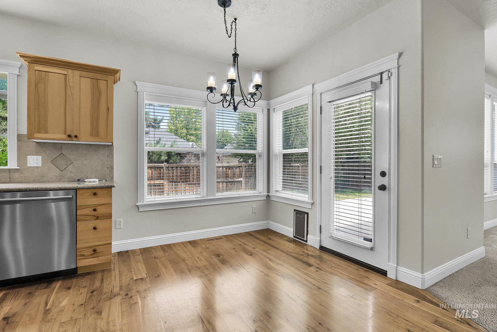 2172 West Cedar Grove Street Meridian, ID 83646 - Photo 12 of 49 Dining area featuring wood-finish flooring, a contemporary chandelier, multiple windows with blinds, a glass-paned door, and light wood cabinetry with a tile backsplash