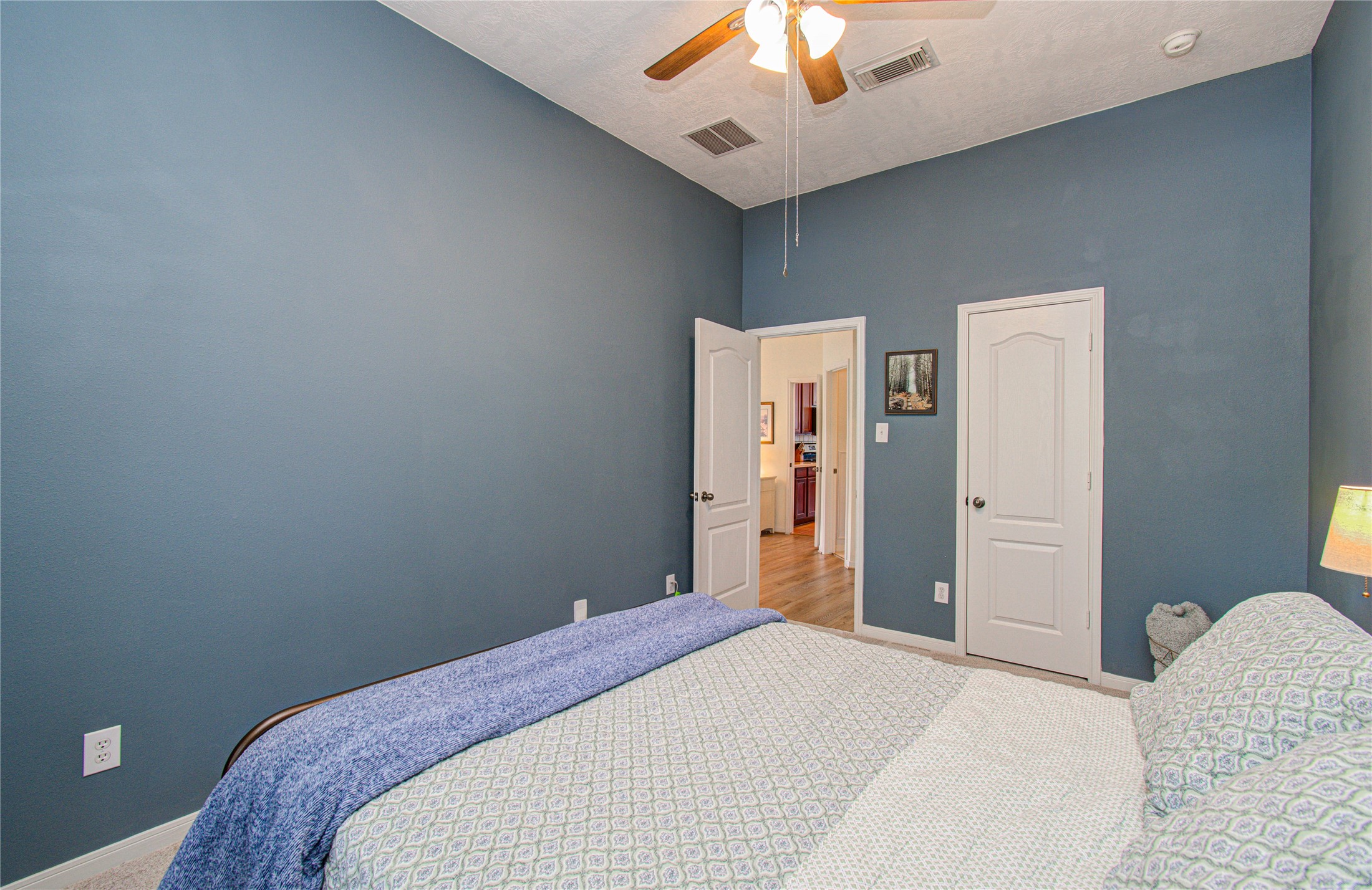 20007 Alcea Court Spring, TX 77379 - Photo 25 of 36 This bedroom features soft blue walls, a ceiling fan, and a cozy bed with patterned bedding. It has two doors, one leading to a hallway with wooden flooring. The room is well-lit and inviting.