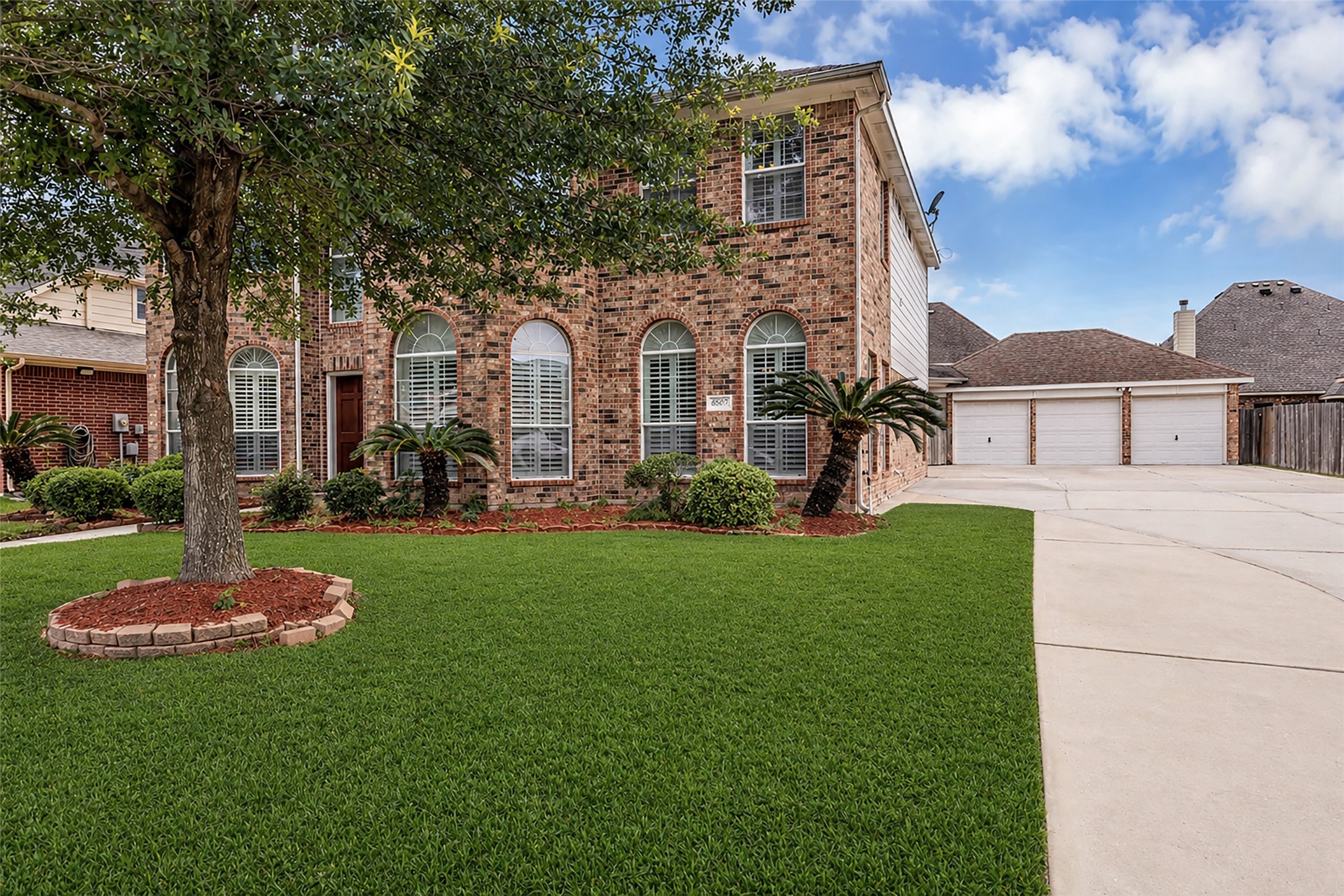 6807 Chapelfield Lane Houston, TX 77049 - Photo 3 of 42 This photo showcases a two-story brick home with a well-maintained front yard. The house features arched windows with shutters and a spacious driveway leading to a three-car garage. A large tree and manicured landscaping add to the curb appeal.