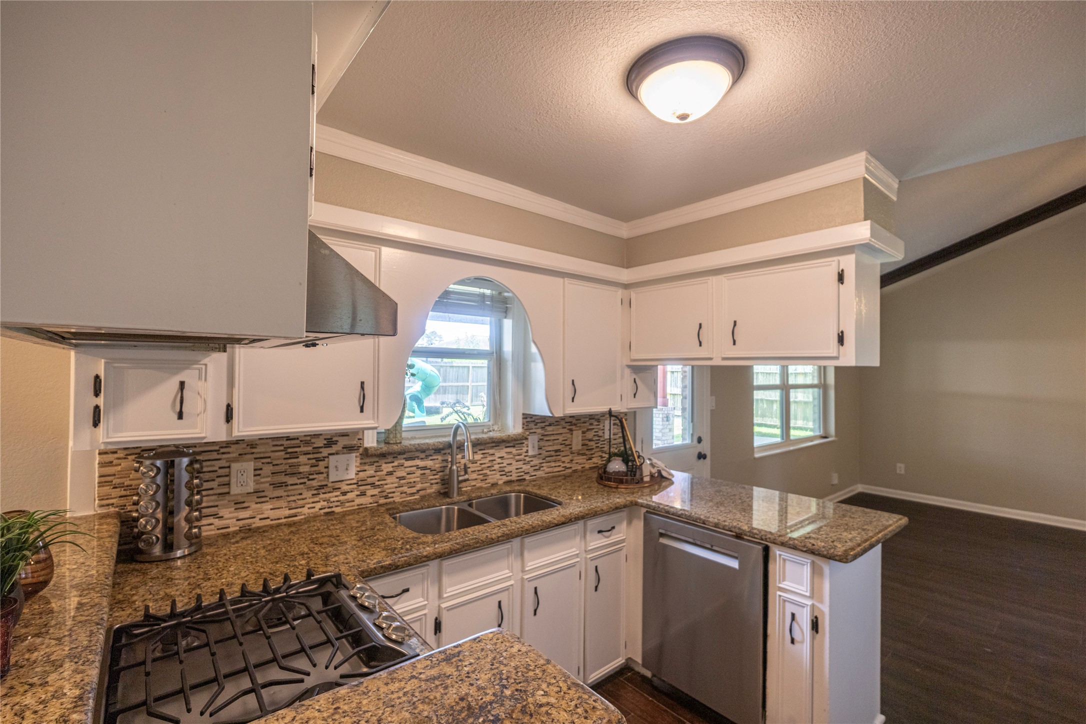 14419 Wadebridge Way Houston, TX 77015 - Photo 4 of 20 This kitchen features granite countertops, a gas range, and white cabinetry with modern hardware. It has a stylish tile backsplash, a double sink under a window, and ample natural light. The open layout connects to the living area, enhancing the spacious feel.