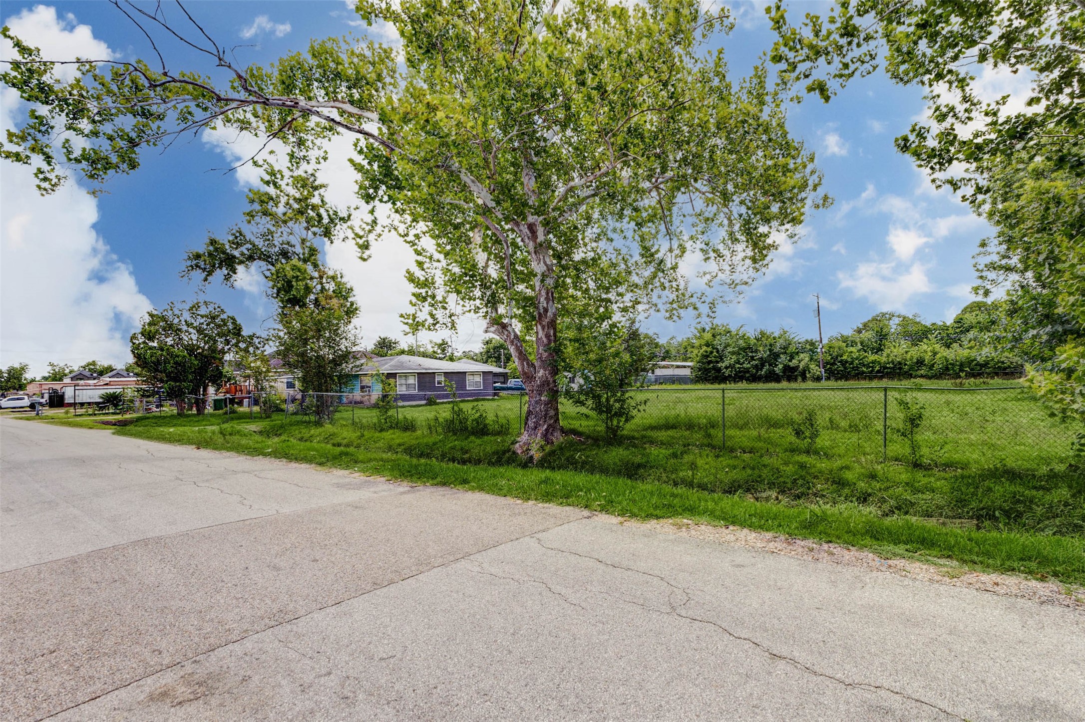 8537 Rannie Road Houston, TX 77080 - Photo 9 of 15 Street-facing view showcasing the property’s frontage, chain-link fencing, mature shade tree, and broad open side yard. This image helps communicate the lot’s width, visibility, and street access, which are key features for land-focused buyers. The large fenced area may appeal to investors, builders, contractors, or users evaluating parking, storage, access, or future development possibilities.