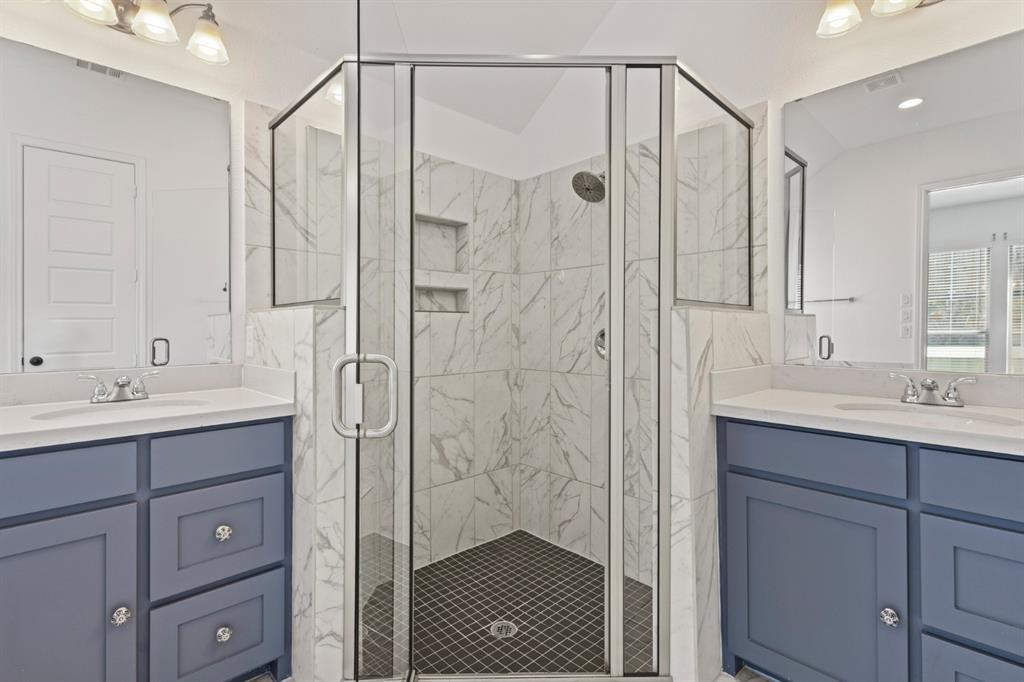 960 Knoxbridge Road Forney, TX 75126 - Photo 13 of 22 Corner glass-enclosed shower with white marble-look tile walls and dark mosaic tile flooring, flanked by dual vanities with blue cabinetry, white countertops, and integrated sinks