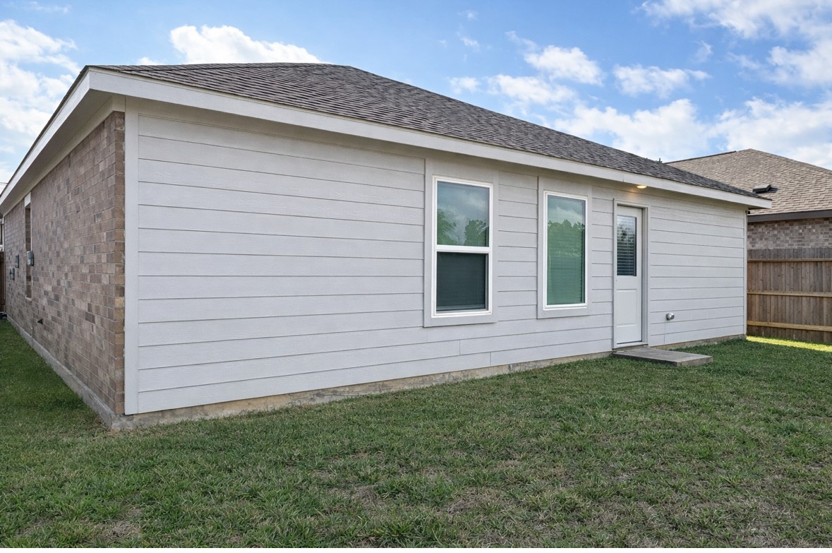 21151 Vercelli Street New Caney, TX 77357 - Photo 15 of 16 This photo shows the back exterior of a single-story home with a combination of brick and siding. It features two windows and a door leading to a grassy backyard, enclosed by a wooden fence. A small concrete step is present at the door.
