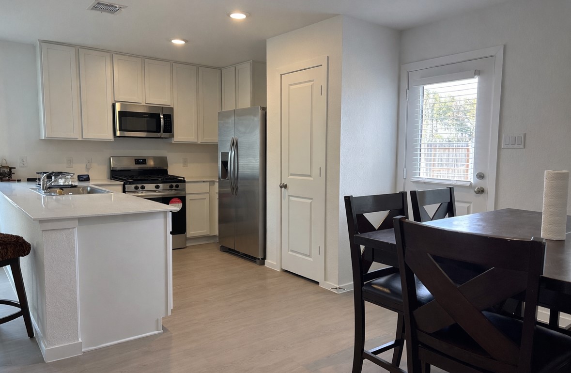 21151 Vercelli Street New Caney, TX 77357 - Photo 4 of 16 This modern kitchen features white cabinetry, stainless steel appliances, and a gas stove. An island with a sink provides extra workspace, while a dining area offers seating. Natural light streams in through a door with vertical blinds, creating a bright, inviting space.