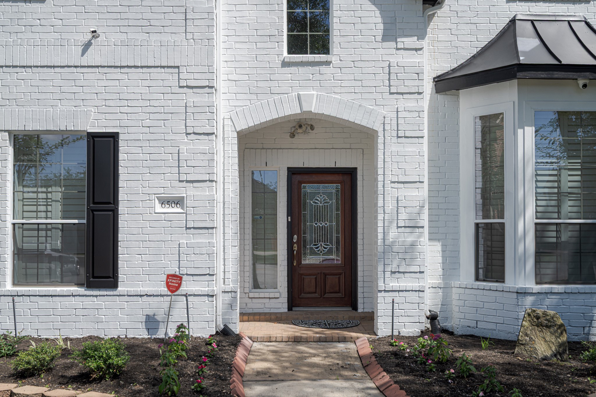 6506 Monte Bello Ridge Lane Houston, TX 77041 - Photo 39 of 39 This photo showcases the entrance of a charming house with a white brick exterior. It features a decorative wooden front door with glass panels, flanked by large windows and a small garden with fresh mulch and plants, creating an inviting and elegant curb appeal.