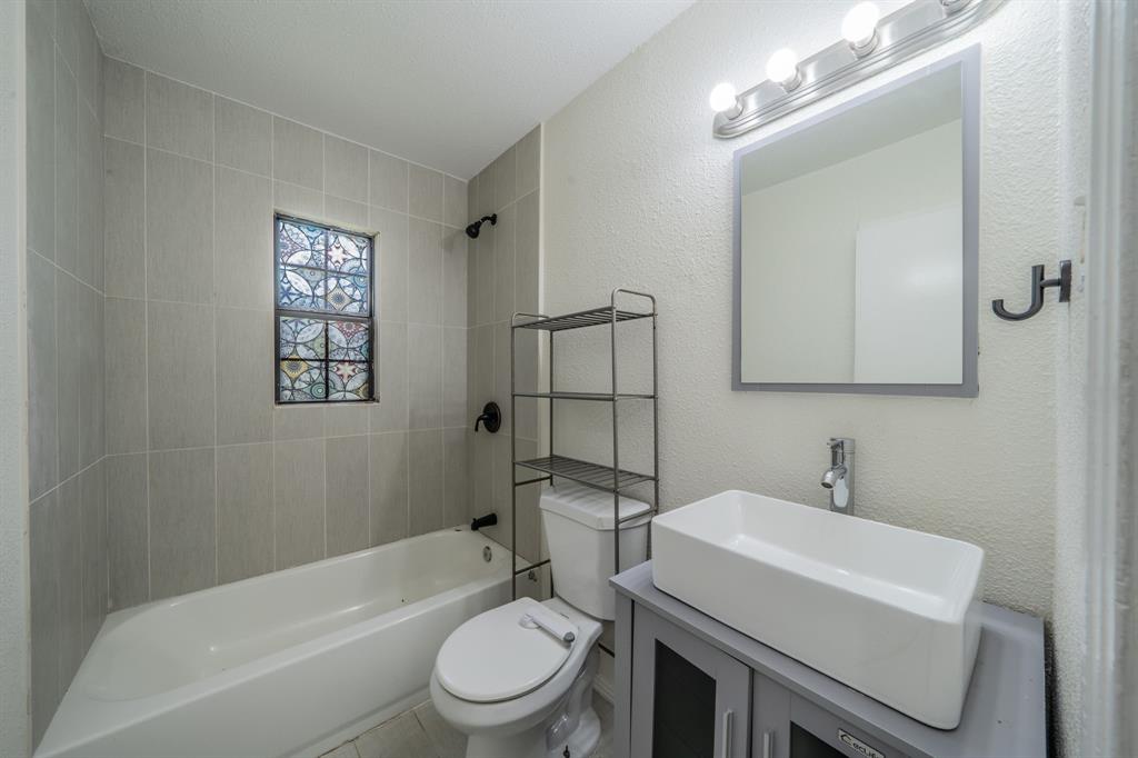 11528 Sinclair Avenue Dallas, TX 75218 - Photo 11 of 16 Bathroom featuring a white bathtub, gray tiled shower surround with a decorative stained glass window, a white vessel sink on a gray vanity, and a wall-mounted mirror with vanity lighting