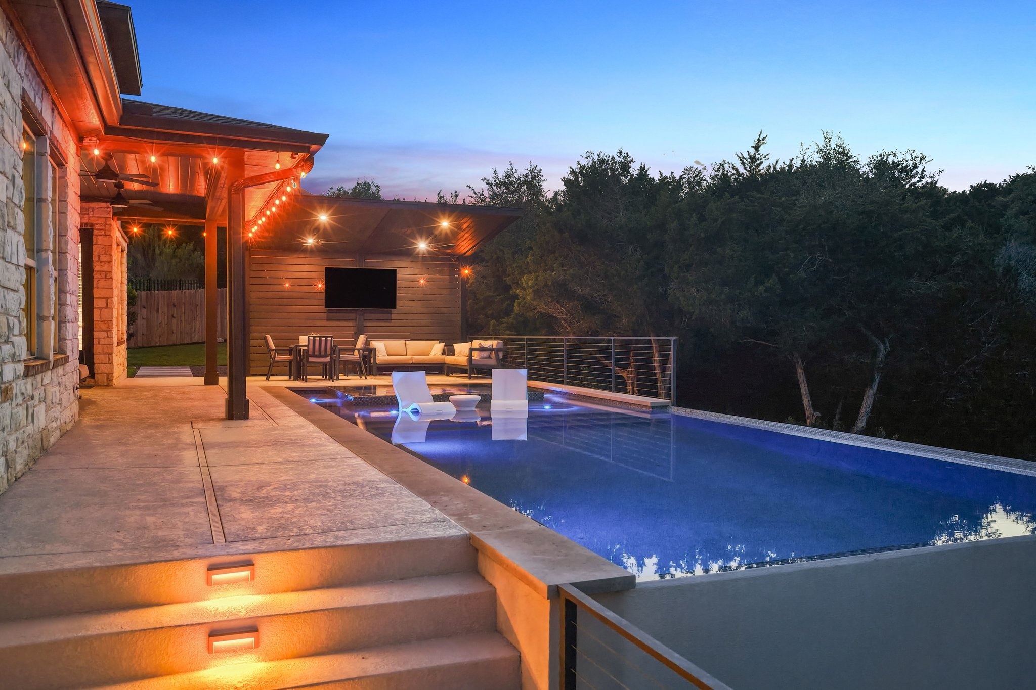 22524 Rock Wren Road Spicewood, TX 78669 - Photo 38 of 40 Outdoor living area featuring a rectangular pool with integrated lighting, a stone exterior, a covered patio with string lighting, a second covered patio with a built-in television, and concrete steps with integrated lighting