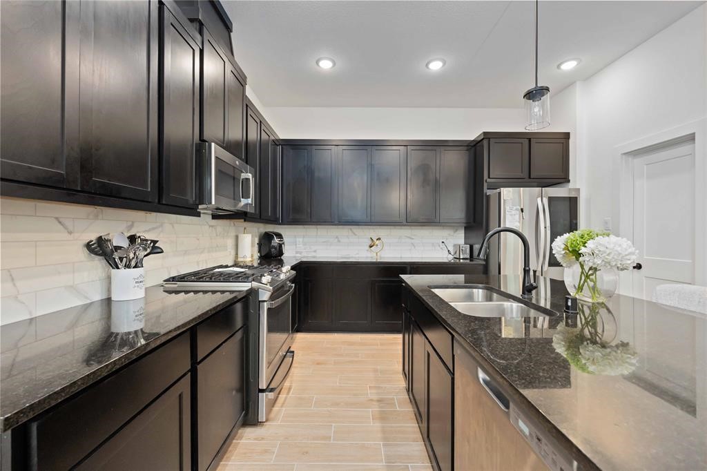 10014 Nectar Path Iowa Colony, TX 77583 - Photo 9 of 26 This modern kitchen features sleek dark cabinetry, stainless steel appliances, and a stylish tile backsplash. The spacious layout includes a large island with a dual sink and ample counter space, perfect for cooking and entertaining.
