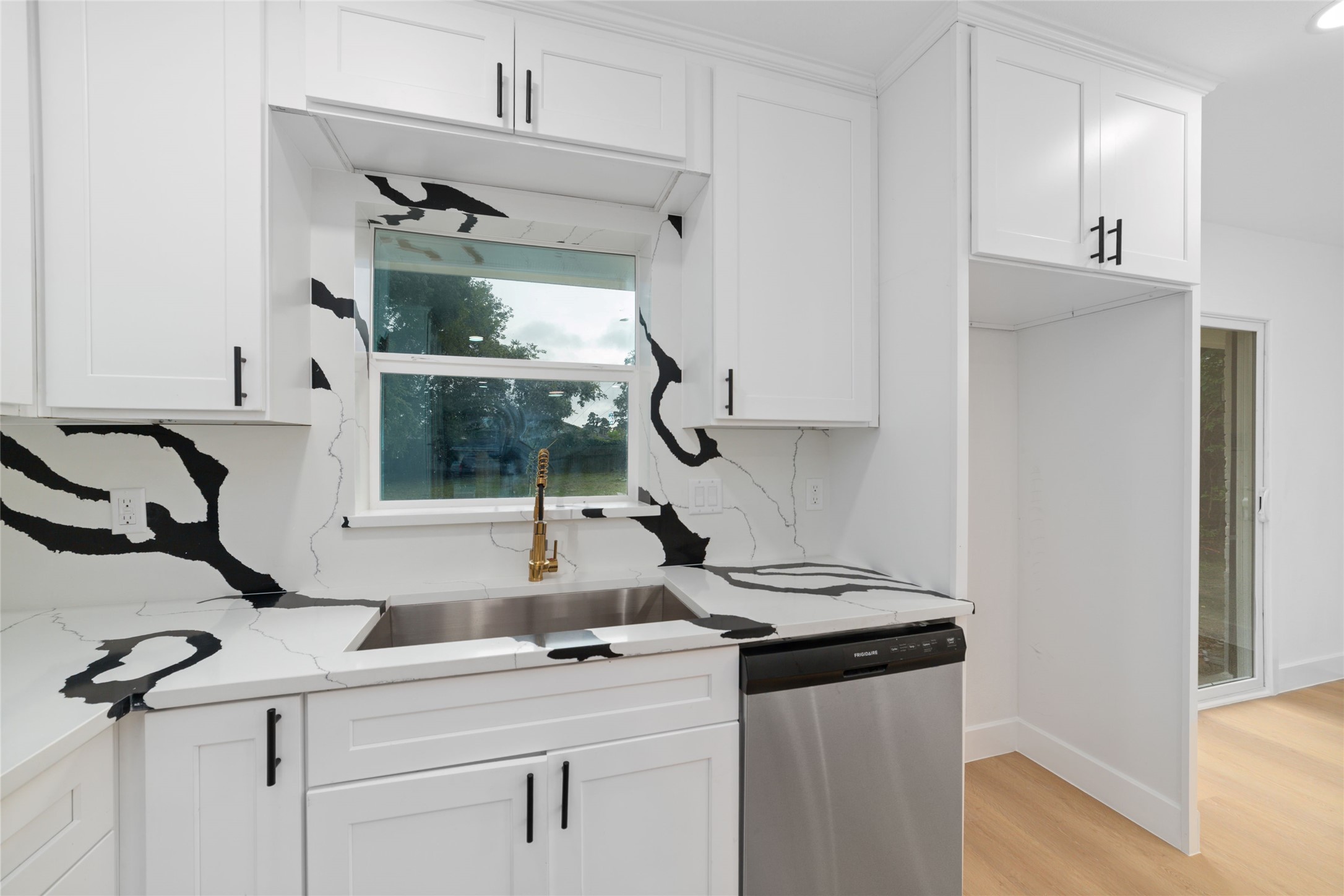 5709 Langley Road Houston, TX 77016 - Photo 9 of 27 This kitchen features modern white cabinetry with black handles, striking black-and-white marble countertops, and a stainless steel dishwasher. A large window above the sink lets in natural light, creating a bright and inviting space.