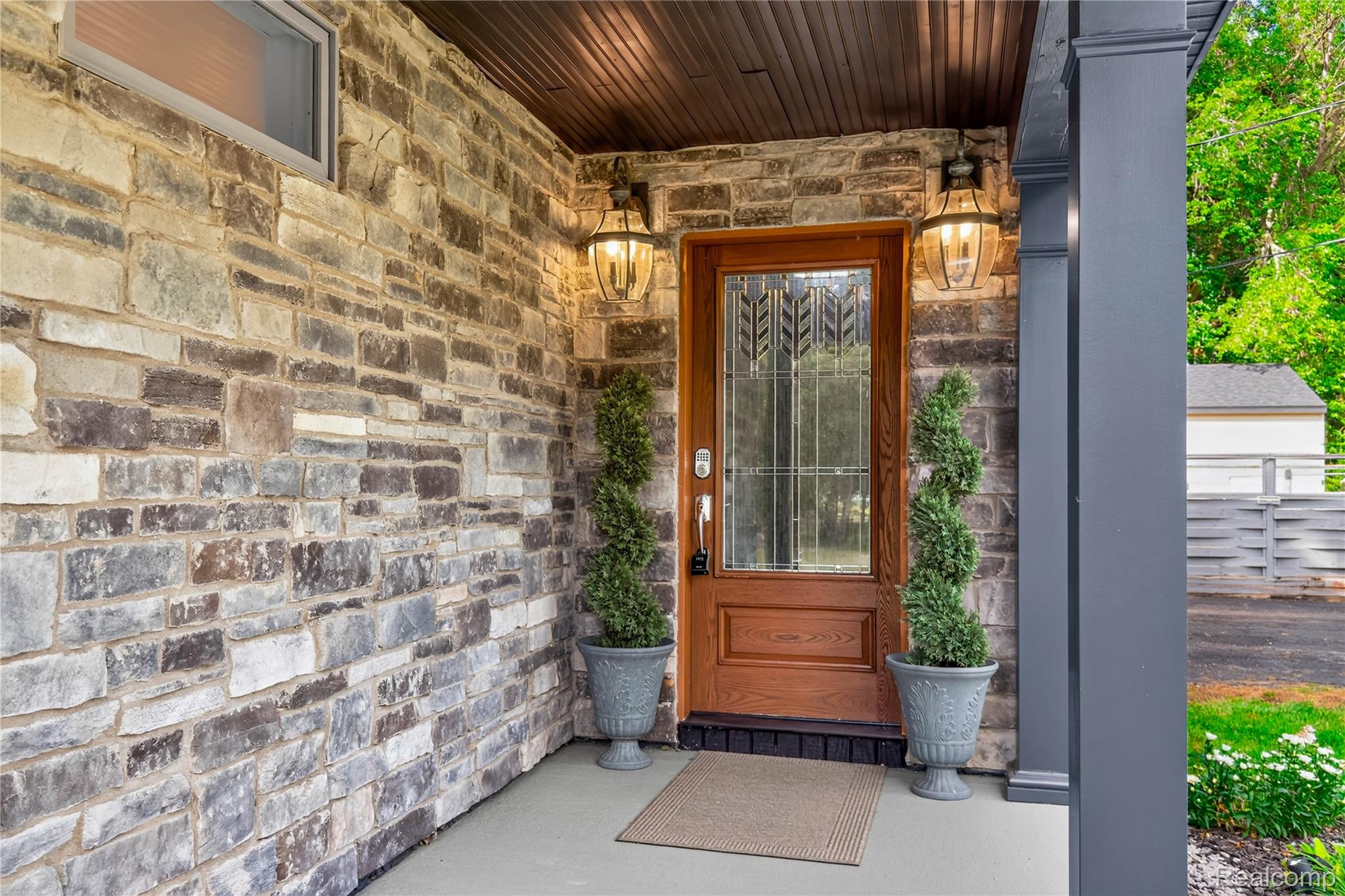4860 Blue Bush Road Monroe, MI 48162 - Photo 4 of 59 Stone veneer exterior featuring a wood-finish front door with decorative glass inserts, an overhead wood-paneled ceiling, and two wall-mounted lanterns