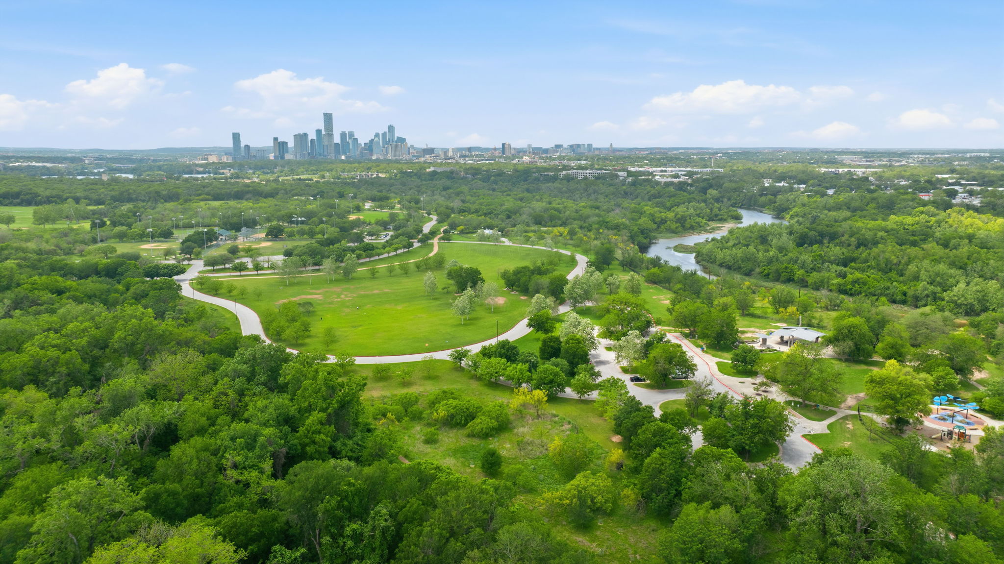 Expansive green spaces and mature trees surround a winding river, with city skyline views visible in the distance