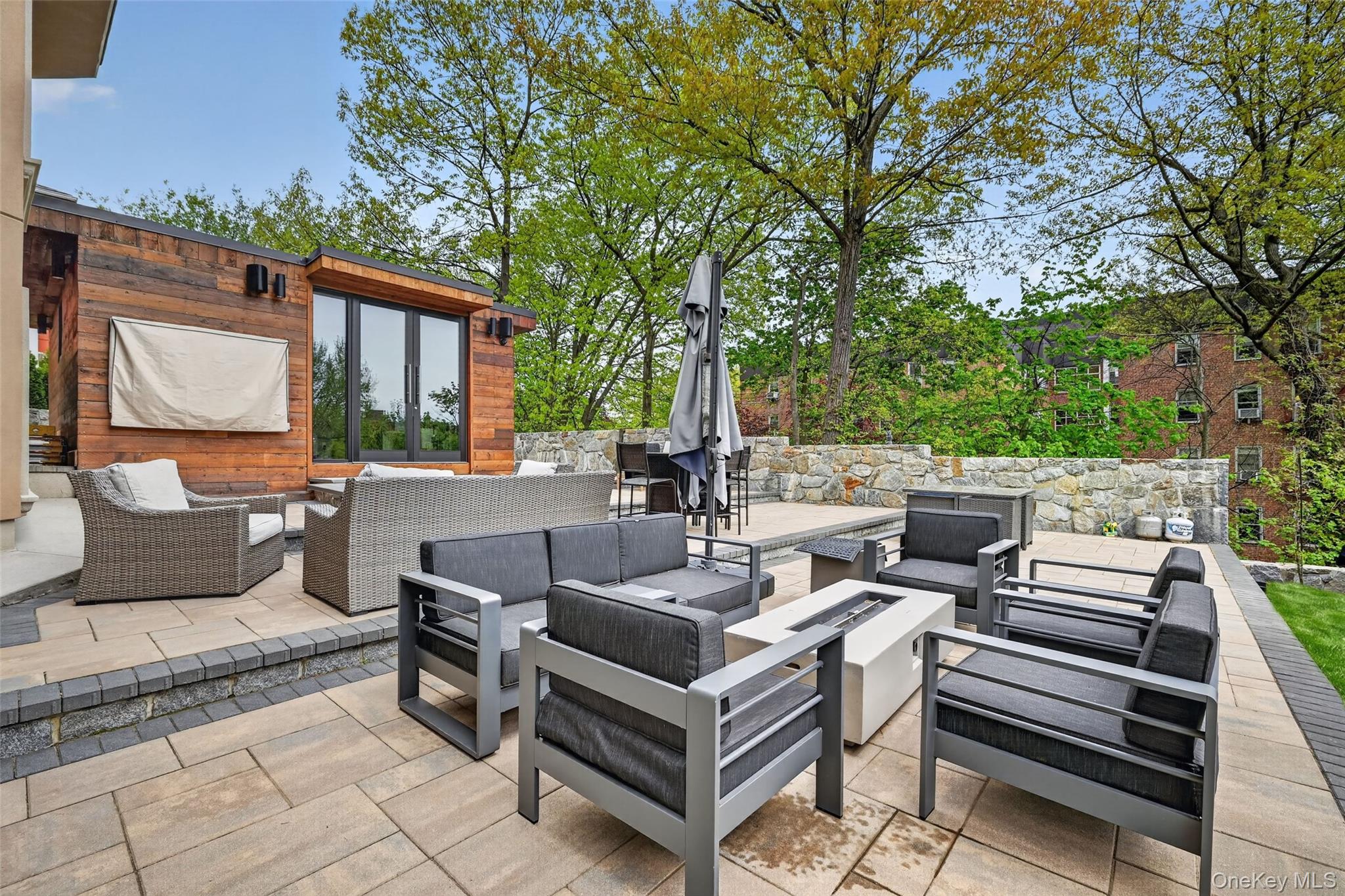 65 Wildway Bronxville, NY 10708 - Photo 21 of 42 Expansive outdoor patio featuring an integrated fire pit, stone retaining walls, and a modern ancillary structure with wood paneling and large glass doors