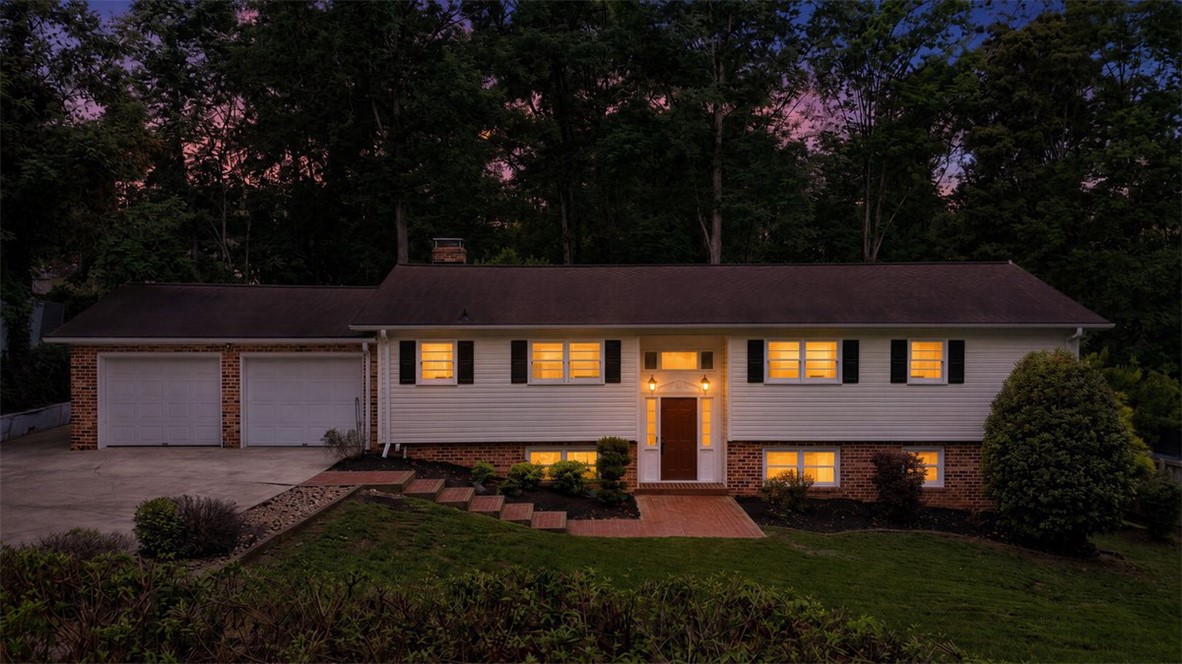 107 Edwards Mill Road Taylors, SC 29687 - Photo 31 of 34 Illuminated windows highlight this inviting split-level home, featuring a two-car garage and a charming brick pathway at twilight.