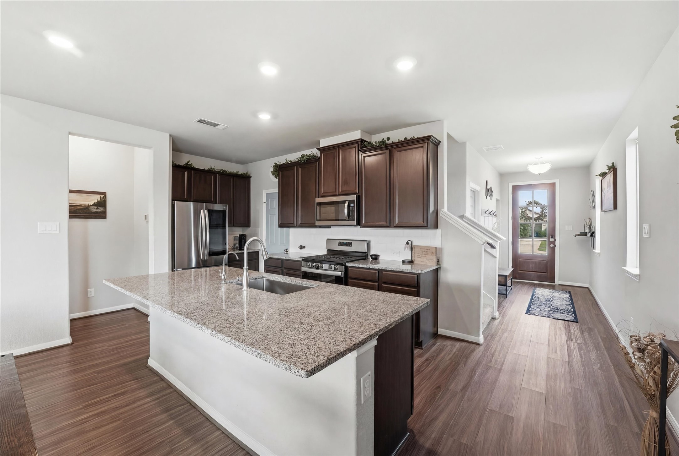 2130 Shellbark Way La Marque, TX 77568 - Photo 5 of 30 This modern kitchen includes a large island with a sink, stainless steel appliances, and dark wood cabinets. The open layout leads to an entryway with a wooden floor and a glass-paneled front door, creating a bright and inviting space.