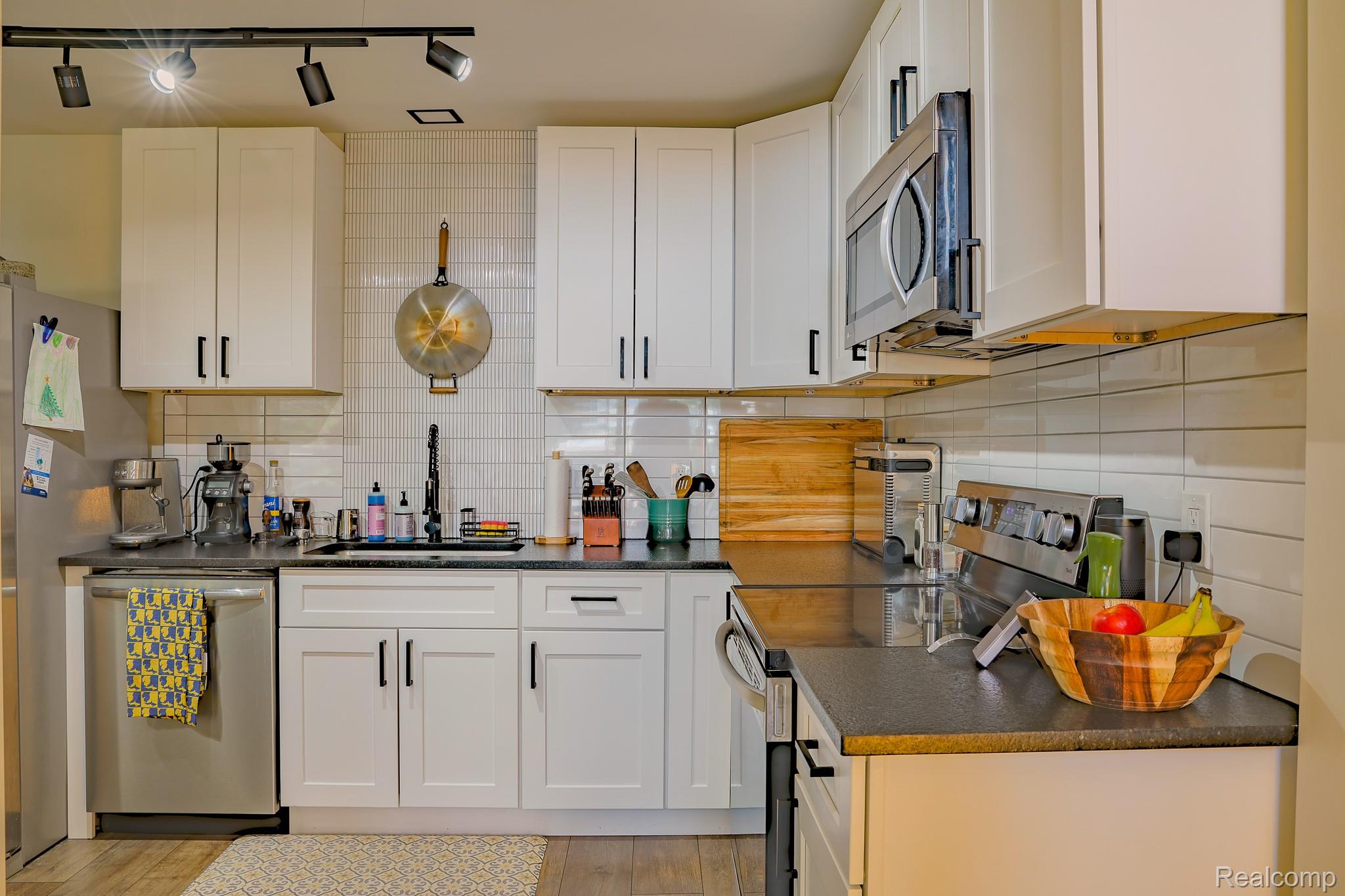 1989 Klingensmith Road, Unit 46 Bloomfield Hills, MI 48302 - Photo 4 of 41 Modern kitchen featuring white shaker-style cabinetry with black hardware, dark countertops, a subway tile backsplash, and stainless steel appliances including a microwave and dishwasher