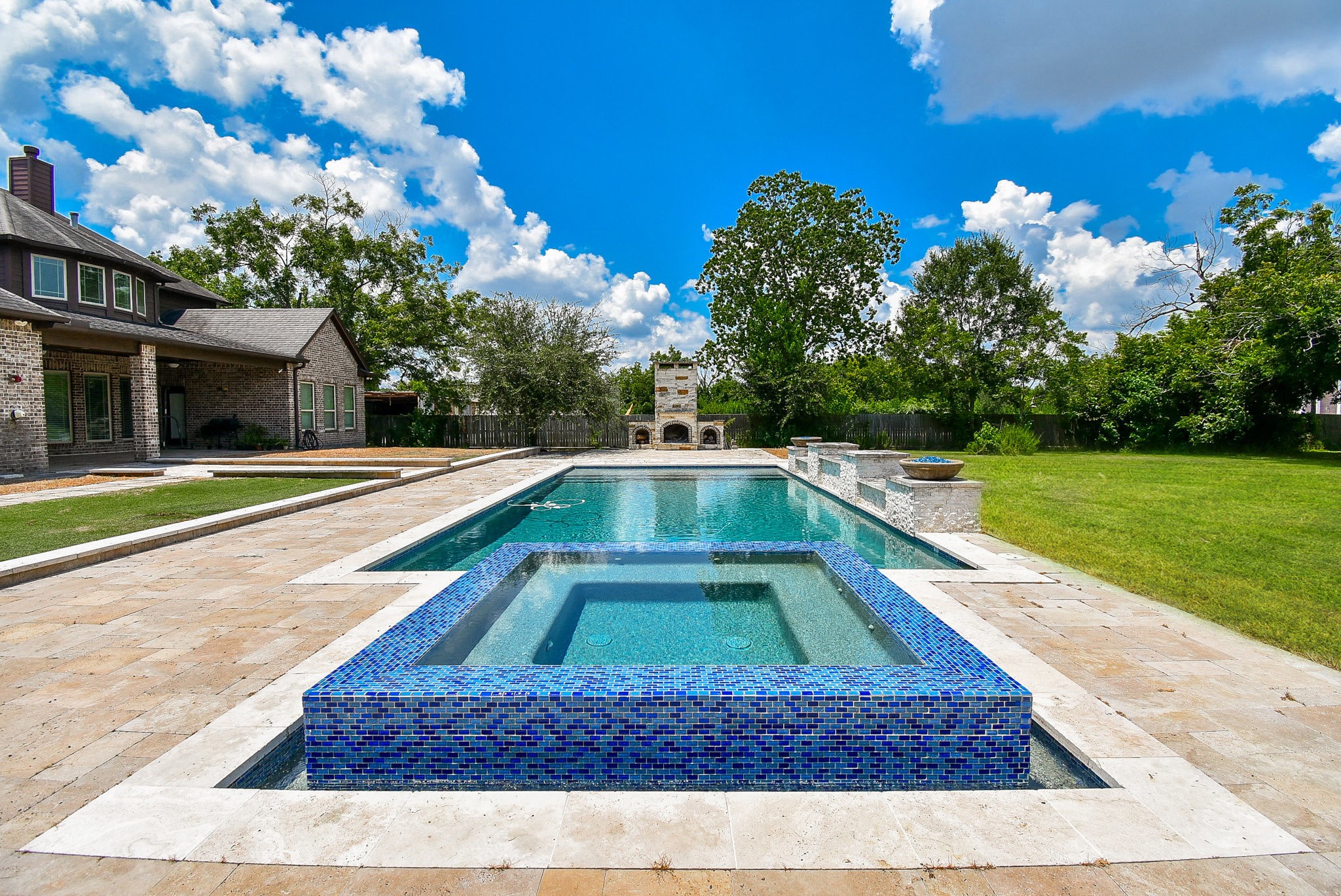 This photo showcases a luxurious backyard with a spacious in-ground pool featuring a hot tub, surrounded by a stone patio. The area includes a large grassy lawn and an outdoor fireplace, perfect for entertaining. The elegant house exterior complements the serene outdoor setting under a bright blue sky.