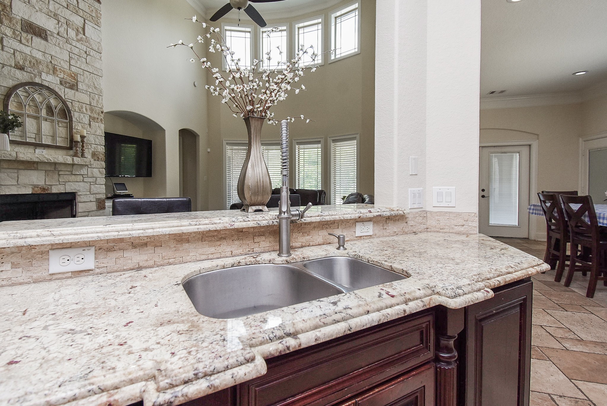 633 Crenshaw Road Houston, TX 77504 - Photo 45 of 46 This photo showcases a modern kitchen with elegant granite countertops and a dual sink. The open floor plan connects to a spacious living area with high ceilings, large windows, and a stone accent wall. The space is bright and airy, ideal for entertaining.