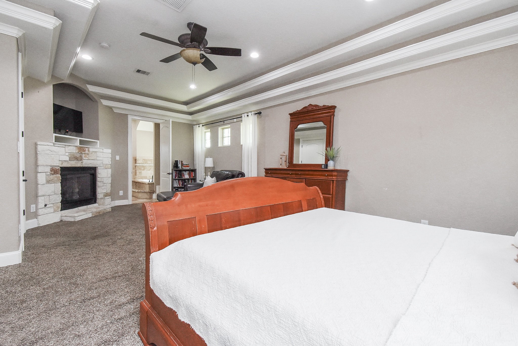 633 Crenshaw Road Houston, TX 77504 - Photo 32 of 46 This spacious bedroom features a cozy stone fireplace, tray ceiling with recessed lighting, and a ceiling fan. It includes a large wooden bed, a dresser with a mirror, and carpeted flooring. Natural light filters through windows, enhancing the room's inviting atmosphere.