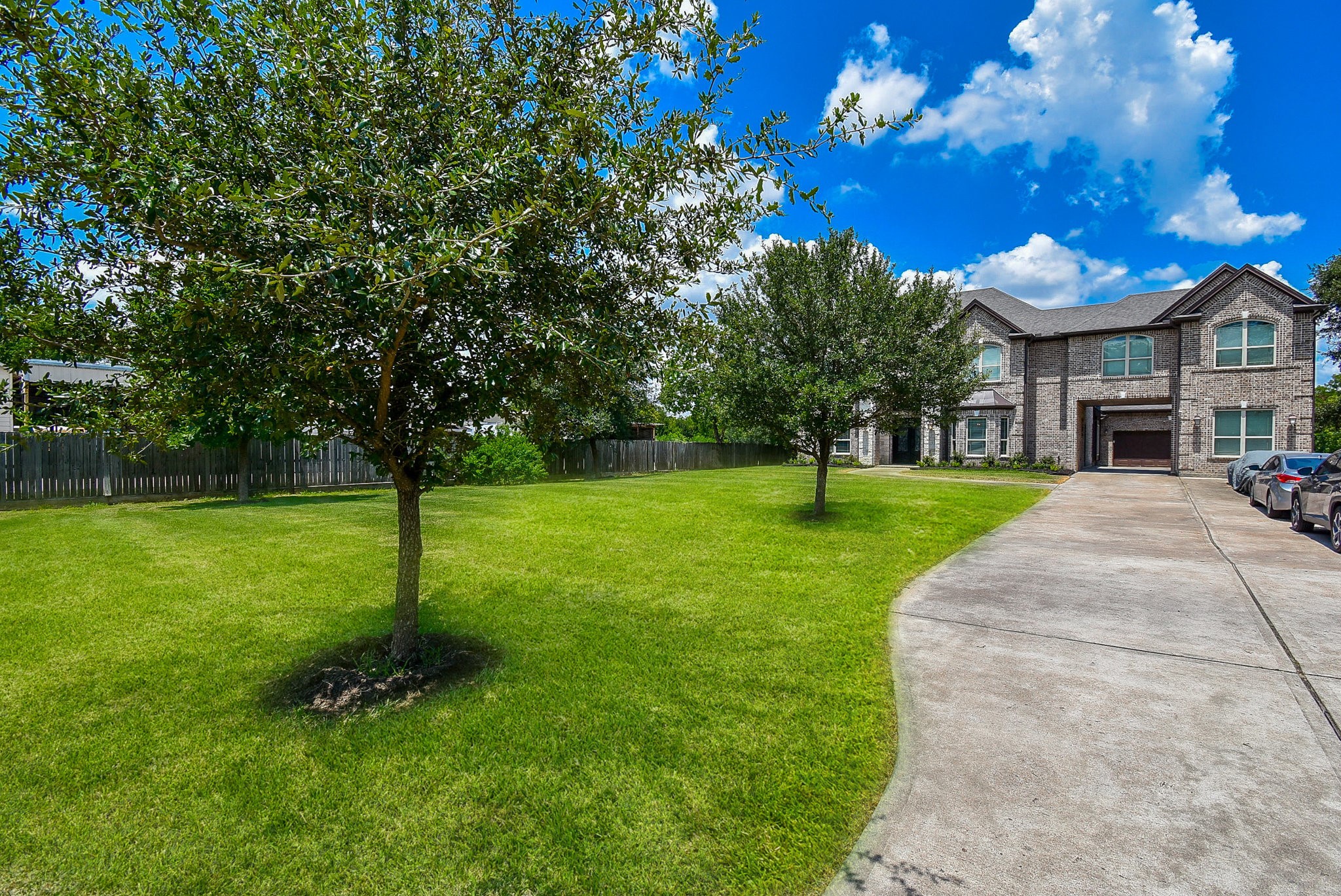633 Crenshaw Road Houston, TX 77504 - Photo 41 of 46 This photo features a spacious two-story brick home with a wide driveway leading to a garage. The house is surrounded by a large, well-maintained lawn with a couple of trees, offering ample outdoor space. The setting is peaceful with a bright blue sky and scattered clouds.