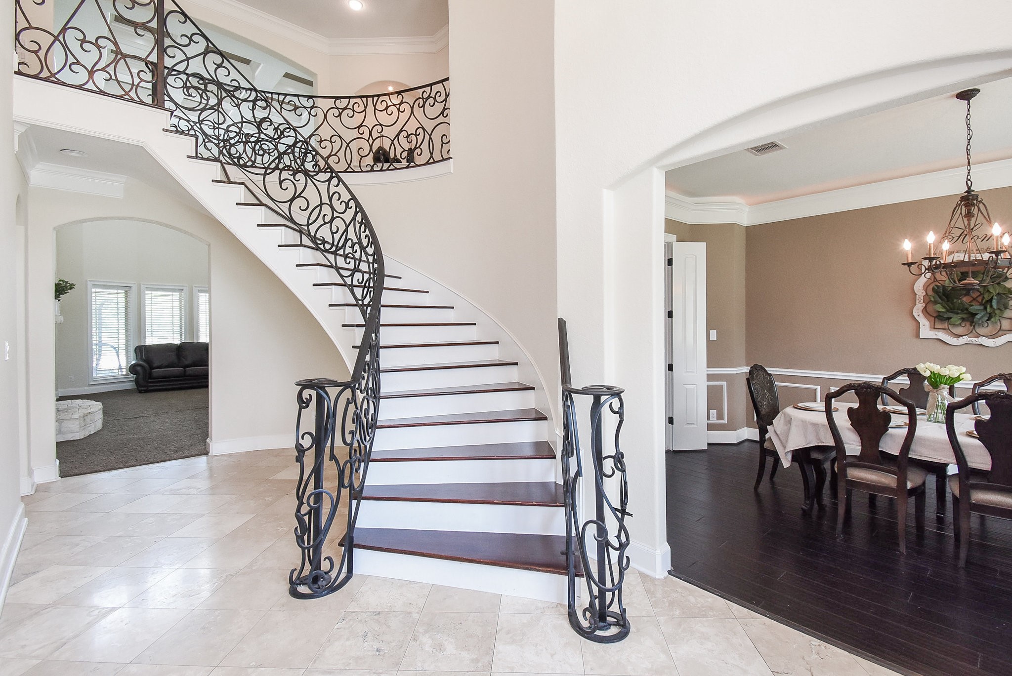 633 Crenshaw Road Houston, TX 77504 - Photo 6 of 46 This elegant home features a grand curved staircase with intricate iron railings, opening into a bright living area with large windows. Adjacent is a formal dining room with dark wood flooring and a chandelier, creating a welcoming atmosphere.