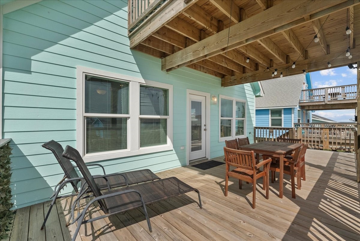130 Beachcomber Avenue Surfside Beach, TX 77541 - Photo 27 of 33 This photo showcases a spacious, sunlit deck with light teal siding, featuring comfortable lounge chairs and a wooden dining set. Overhead string lights add ambiance, and the area is shaded by an upper balcony, perfect for outdoor relaxation and entertaining.