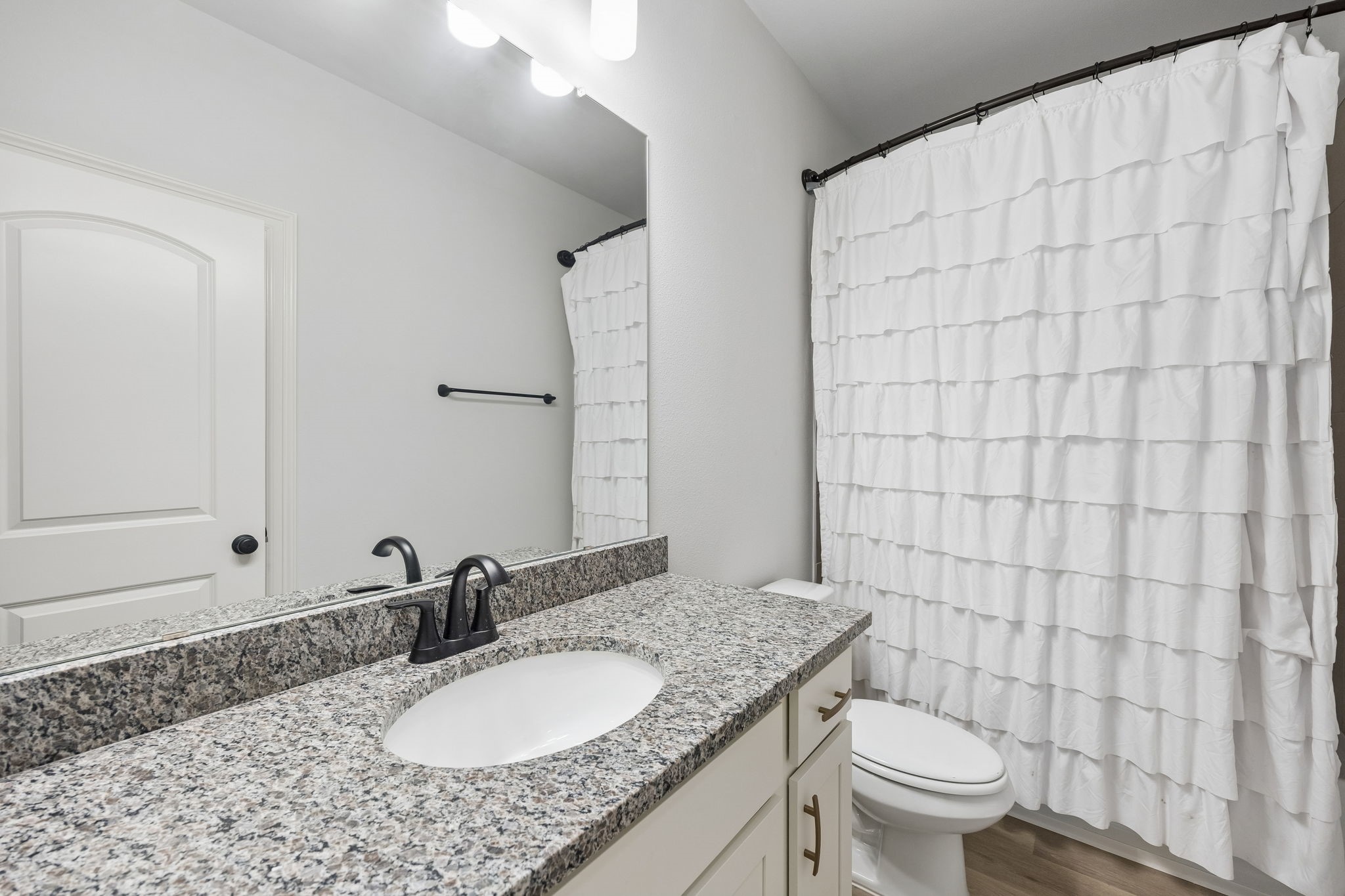 10902 Bonfire Trail Beasley, TX 77417 - Photo 19 of 36 This bathroom features a modern design with a granite countertop, undermount sink, and sleek black fixtures. It includes a large mirror, a white vanity, and a ruffled shower curtain, creating a fresh and clean look.