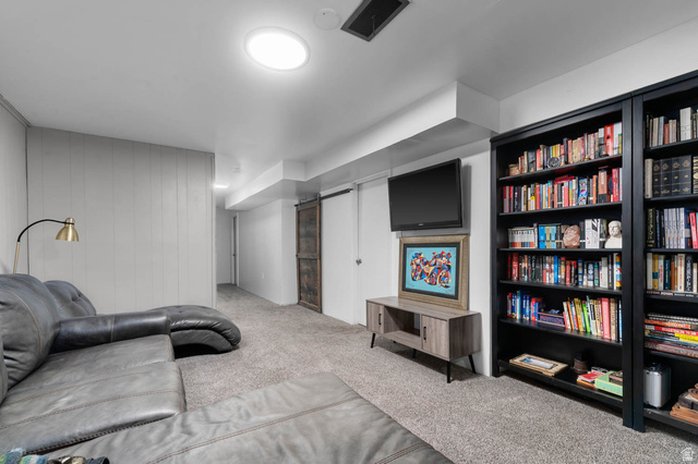 9902 South Poppy Lane Sandy, UT 84094 - Photo 17 of 25 Carpeted living area featuring a wood-panel accent wall, built-in black shelving units, a ceiling-mounted light fixture, and a wood-finish sliding barn door
