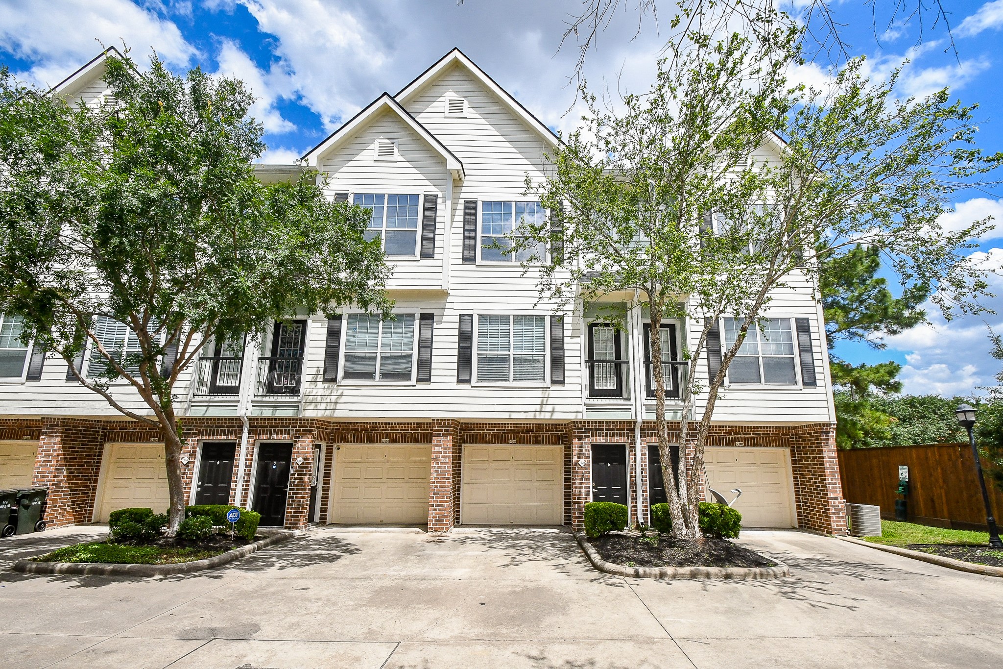 This is a modern, multi-story townhouse with a brick and siding exterior. It features garages and front entrances, framed by well-maintained trees and landscaping. The home offers a clean, welcoming facade.