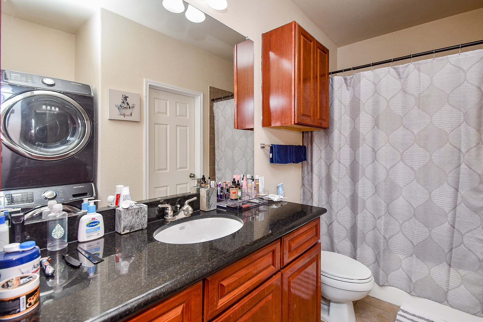 9200 Westheimer Road, Unit 1302 Houston, TX 77063 - Photo 26 of 29 Modern bathroom with a sleek granite countertop, under-mount sink, and warm wooden cabinetry. Features a stacked washer and dryer, shower with a patterned curtain, and ample lighting.