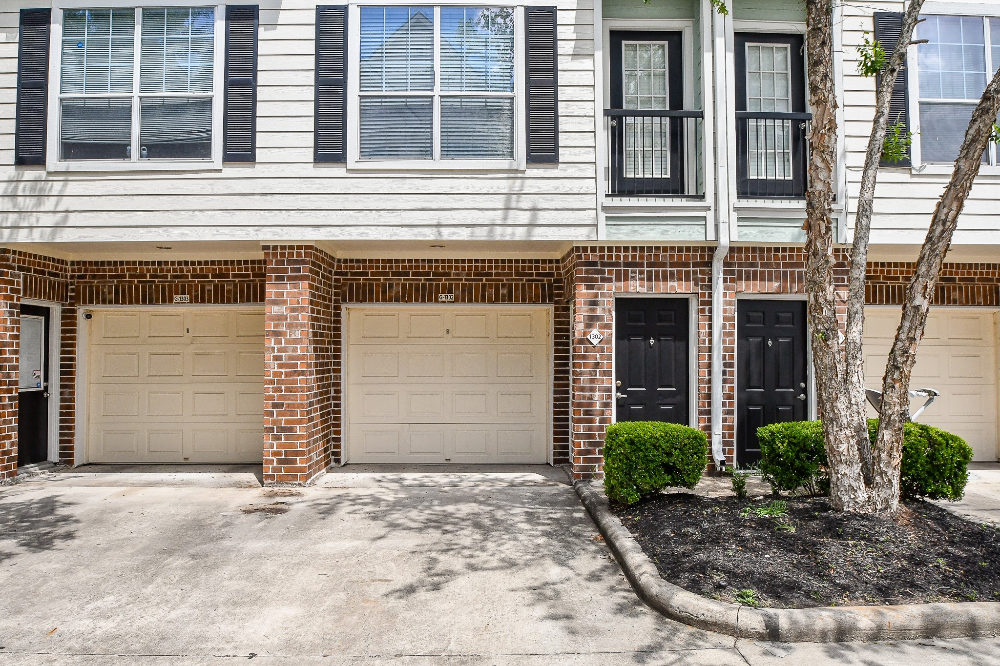 9200 Westheimer Road, Unit 1302 Houston, TX 77063 - Photo 4 of 29 This photo shows a townhouse exterior with brick accents and siding. It features one-car garage, each with a black front door adjacent. The upper level has windows with shutters, and there are small shrubs and trees in front.
