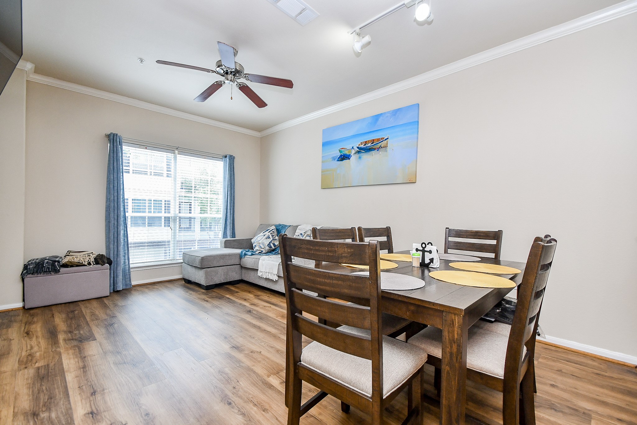 9200 Westheimer Road, Unit 1302 Houston, TX 77063 - Photo 10 of 29 This photo shows a bright, open-concept living and dining area with wood flooring. It features a cozy gray sectional near a large window with blue curtains, providing plenty of natural light. The dining area has a wooden table set for six, and a ceiling fan and track lighting enhance the space.