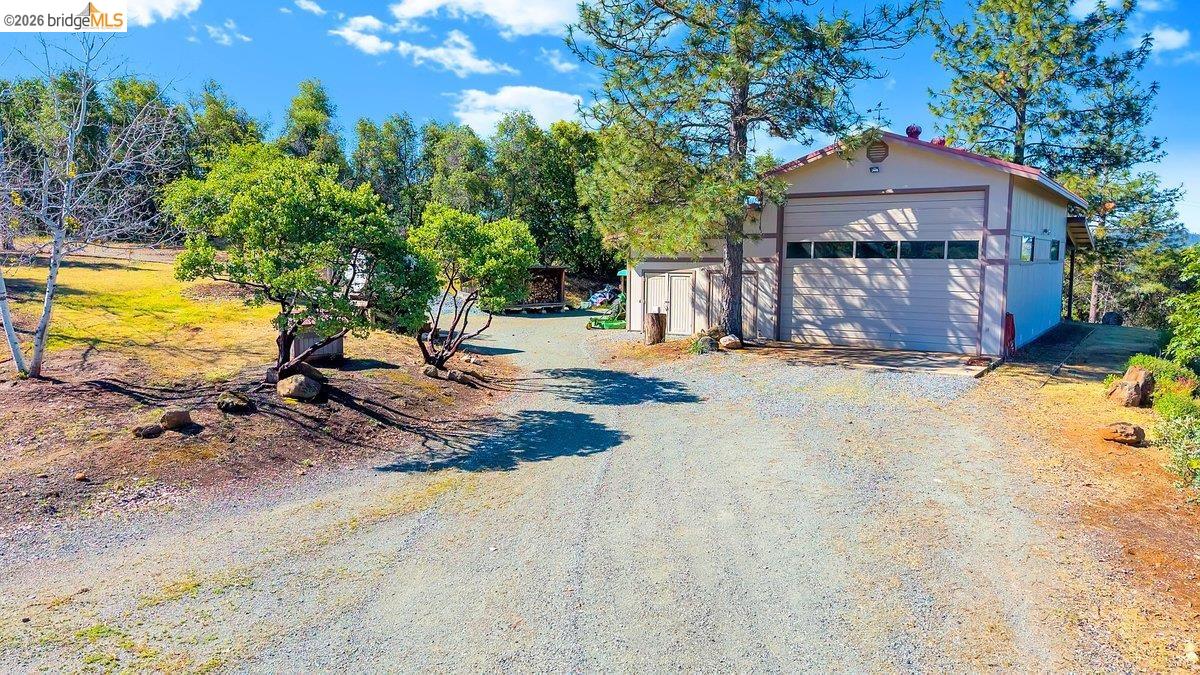 13700 Clements Road Groveland, CA 95321 - Photo 91 of 93 Gravel driveway leading to a detached workshop with a roll-up door, multiple windows, and exterior lighting