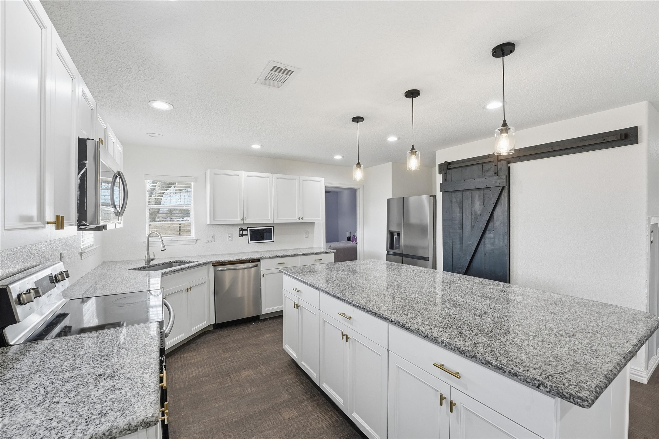 611 Reidland Road Crosby, TX 77532 - Photo 11 of 35 Modern kitchen featuring a large granite island, white cabinetry, and stainless steel appliances. It has a farmhouse-style sliding barn door, pendant lighting, and ample natural light. Perfect for family gatherings and entertaining.