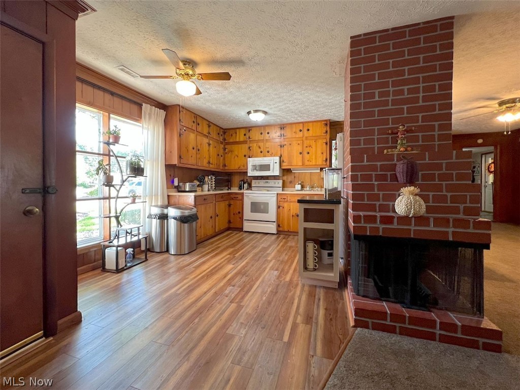 Kitchen featuring wood-finish flooring, wood cabinetry, white appliances, a ceiling fan, and a brick fireplace with a hearth