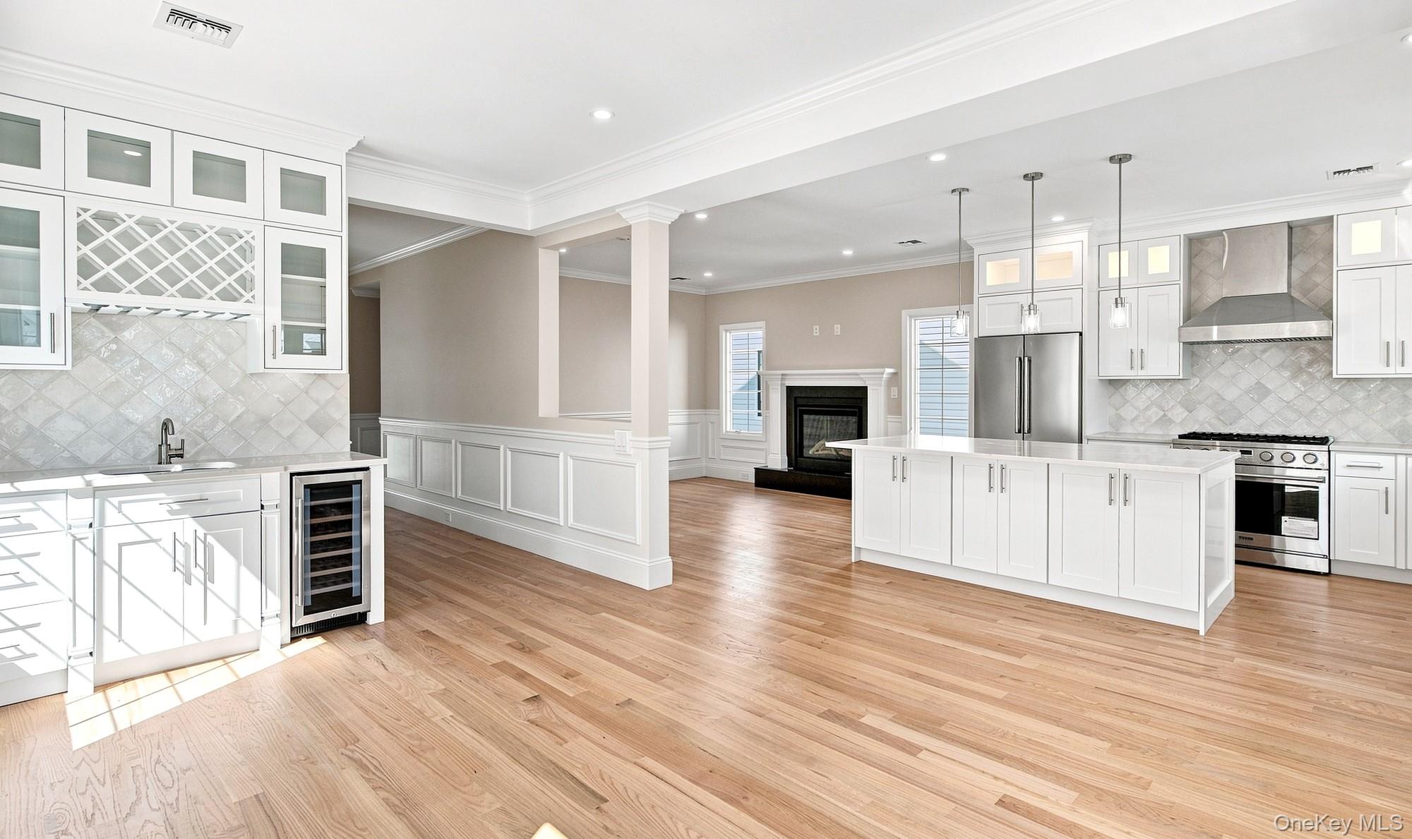 148 Latham Road Mineola, NY 11501 - Photo 10 of 27 Great Room cross-view of kitchen, family room and dining. Light wood-finish flooring throughout, white cabinetry with glass-front uppers, stainless steel appliances, central kitchen island with pendant lighting, and a built-in beverage refrigerator