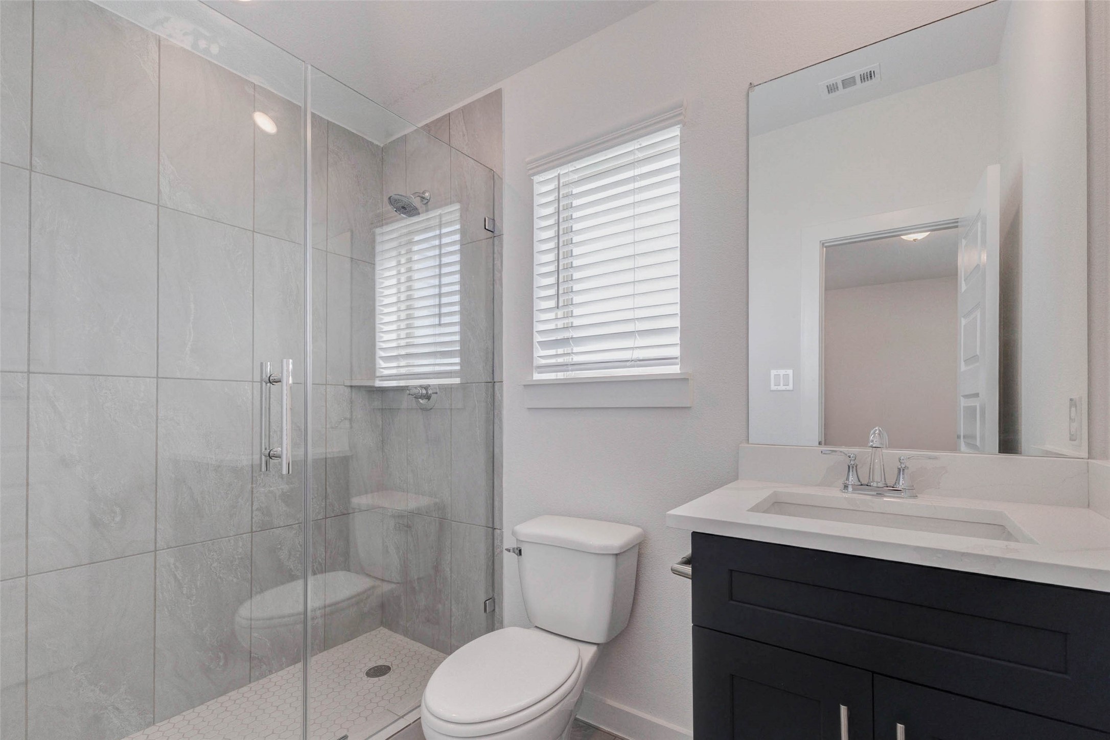1038 Elsinore Drive Rosharon, TX 77583 - Photo 20 of 27 The primary bathroom showcases a sleek and modern design featuring a spacious glass-enclosed walk-in shower with elegant tile surround and contemporary fixtures. A stylish vanity with quartz countertop and dark cabinetry provides both storage and visual contrast, while the large mirror enhances the bright, clean aesthetic. Natural light from the window adds warmth and creates a refreshing spa-like environment for daily routines.