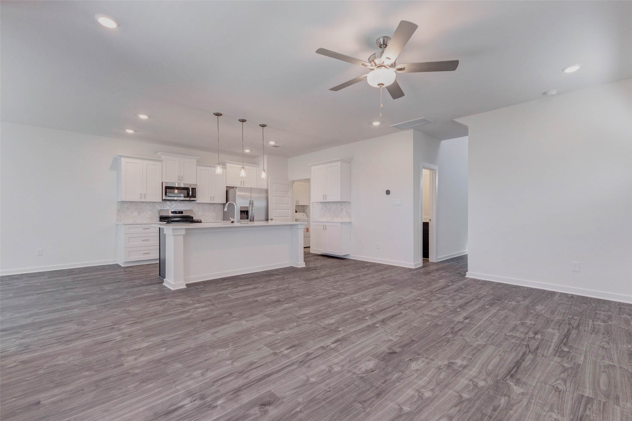 1038 Elsinore Drive Rosharon, TX 77583 - Photo 26 of 27 This wide view of the main living space highlights the seamless connection between the kitchen, dining, and living areas. The open floor plan creates an airy atmosphere perfect for entertaining or relaxing at home. Pendant lighting above the island, recessed ceiling lights, and a modern ceiling fan complement the bright neutral color palette, while the expansive layout offers flexibility for a variety of furniture arrangements.