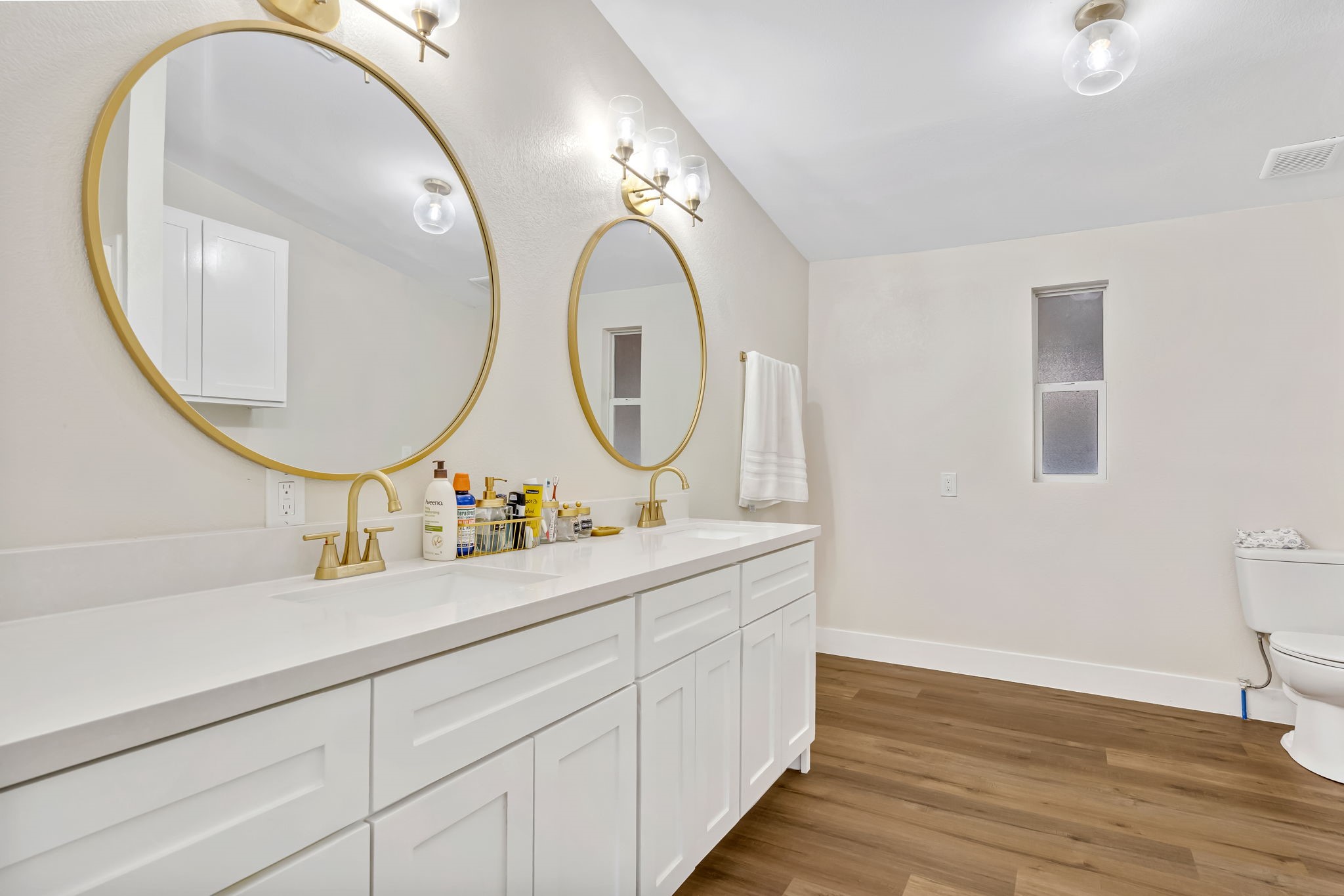 1491 Pecan Valley Road Wharton, TX 77488 - Photo 15 of 38 This master bathroom boasts a sleek double vanity with white cabinetry and elegant countertops. Gold-accented mirrors and matching fixtures enhance the sophisticated style, while wood-style flooring and a frosted window create a bright, airy, and private space.