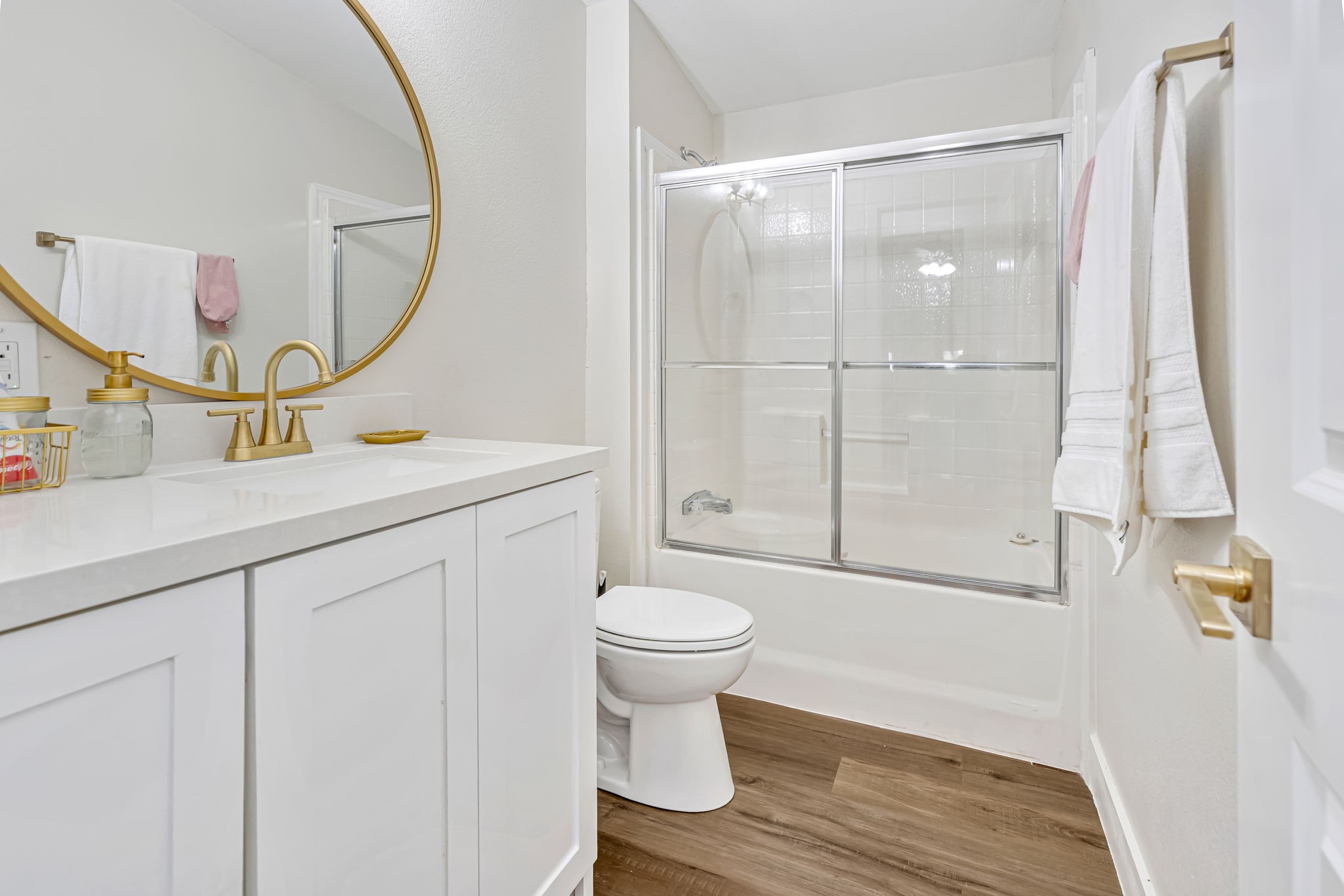 1491 Pecan Valley Road Wharton, TX 77488 - Photo 22 of 38 Experience modern elegance in this bathroom, featuring a large round mirror, gold fixtures, and a sleek white vanity. The shower-tub combo with sliding glass doors offers a clean and functional space.