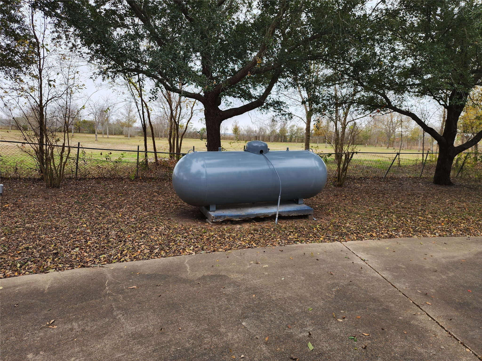 1491 Pecan Valley Road Wharton, TX 77488 - Photo 28 of 38 A spacious backyard features a large propane tank, nestled among trees and enclosed by a chain-link fence, offering a natural, private setting.