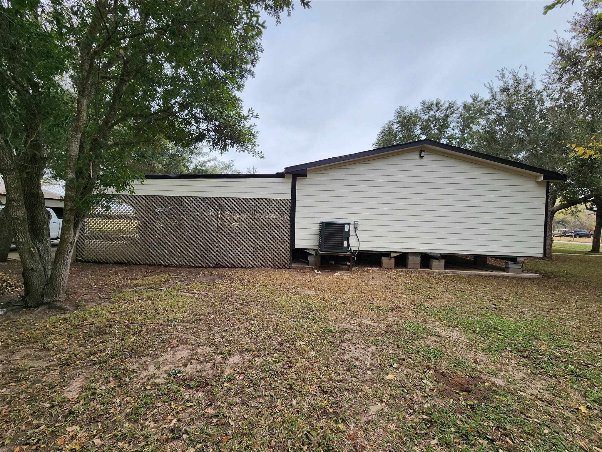 1491 Pecan Valley Road Wharton, TX 77488 - Photo 3 of 38 Charming manufactured home featuring light-colored siding and a stylish lattice panel, elevated for a modern touch. Nestled in a lush grassy yard with mature trees, it includes an exterior air conditioning unit for year-round comfort.