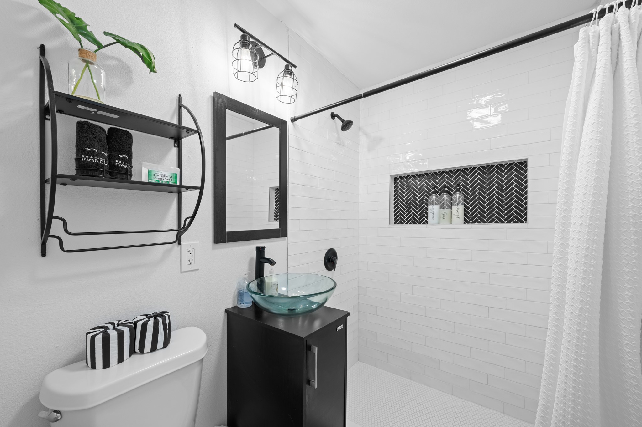 729 Kyle Angleton, TX 77515 - Photo 27 of 36 This bathroom features a modern design with sleek white tiles, a glass vessel sink, and a black vanity. There's a stylish shower area with a herringbone accent and a functional shelving unit. The space is bright and contemporary.