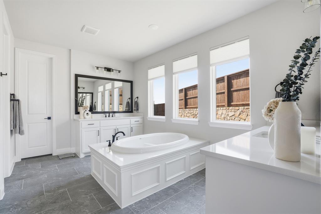 4202 Crown Run Road Midlothian, TX 76065 - Photo 14 of 34 Spacious bathroom featuring a built-in soaking tub with paneled surround, three large windows, a double vanity with white cabinetry and dark hardware, and grey tile flooring