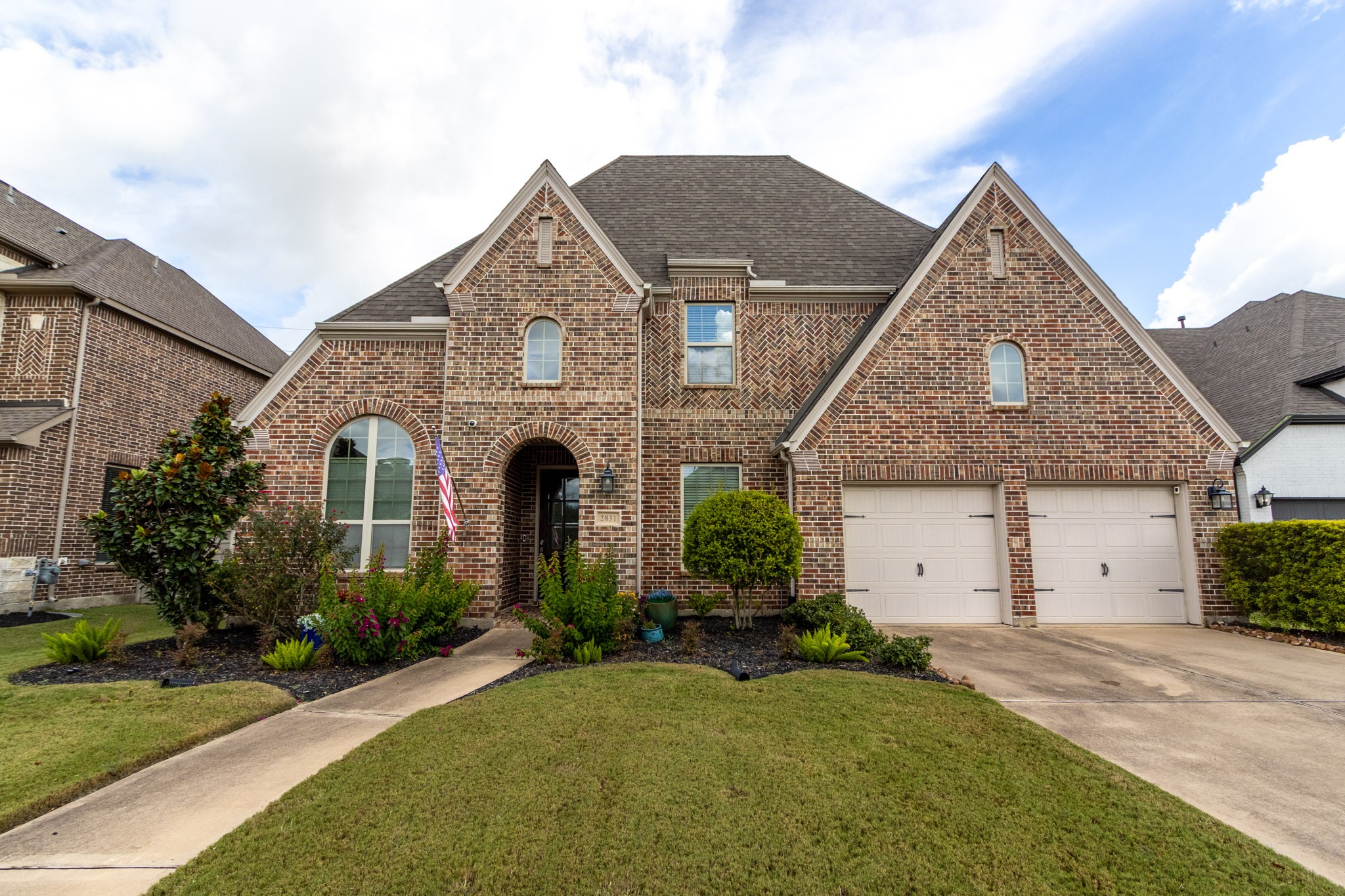 This charming brick home features a classic design with a steep roof and arched windows. It includes a spacious two-car garage, a well-maintained front yard with tasteful landscaping, and a welcoming walkway leading to the entrance.