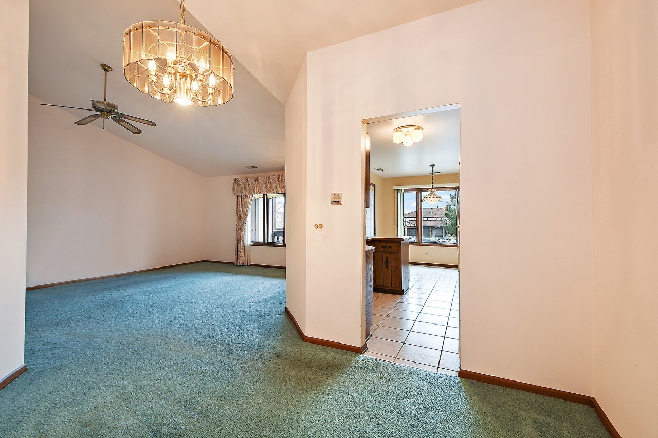 9722 West Creek Road, Unit E2 Palos Park, IL 60464 - Photo 3 of 18 an empty room with a chandelier fan and a kitchen view