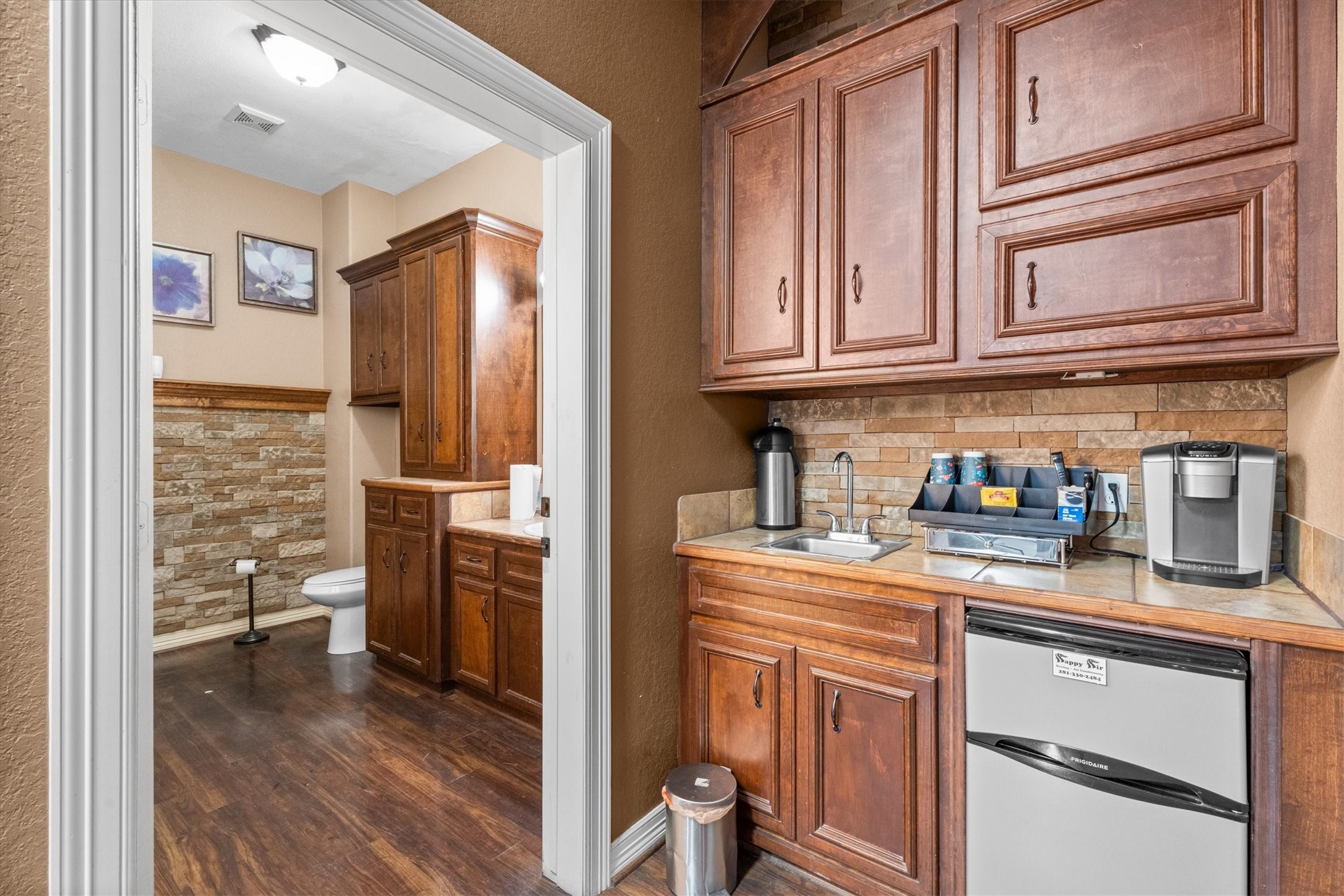11066 Sh 242 Conroe, TX 77385 - Photo 11 of 21 This photo shows a compact kitchenette area with wooden cabinets, a small sink, a mini-fridge, and a coffee maker. Adjacent is a bathroom with rustic-style stone wall accents, wooden cabinetry, and hardwood flooring.